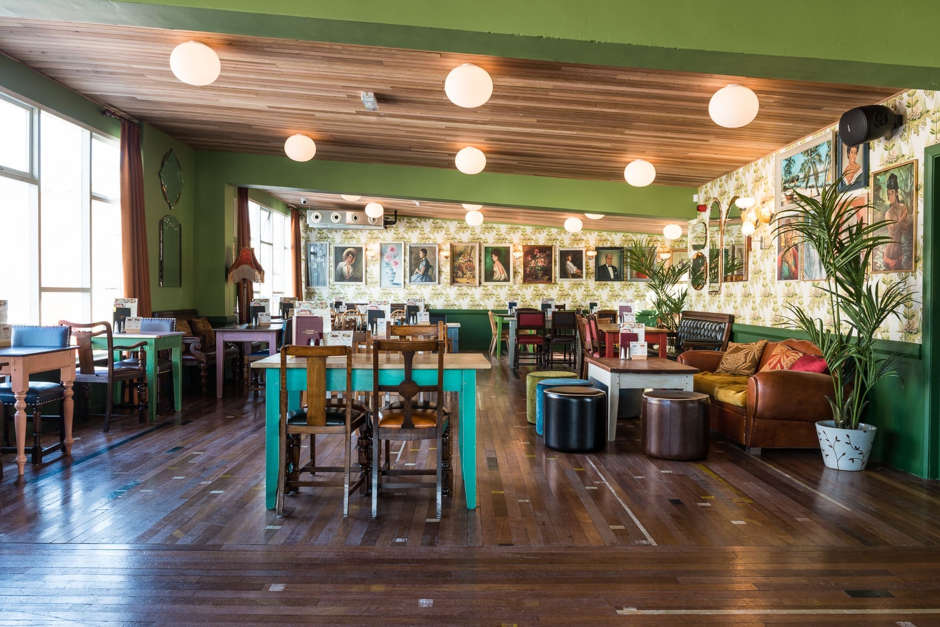 A spacious, sunlit café by Rivo with wooden floors, eclectic furniture, green walls, round hanging lights, potted plants, and framed portraits on a wallpapered wall in the background.