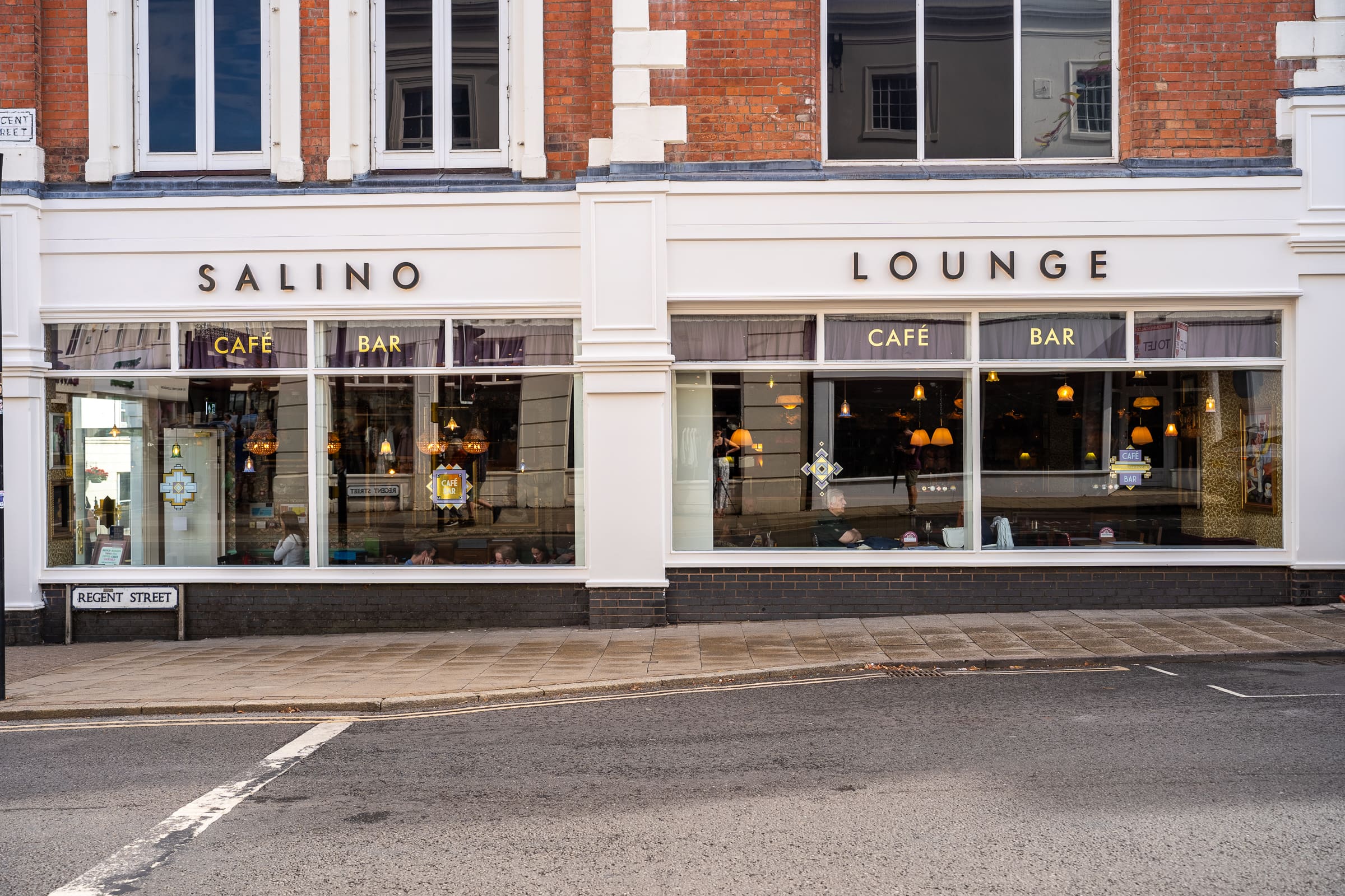 The exterior of Salino Lounge, a vibrant café and bar with large windows, visible seating, and warm hanging lights, is set on a street corner with a sign reading "Regent Street." Enjoy the inviting atmosphere only at Salino.