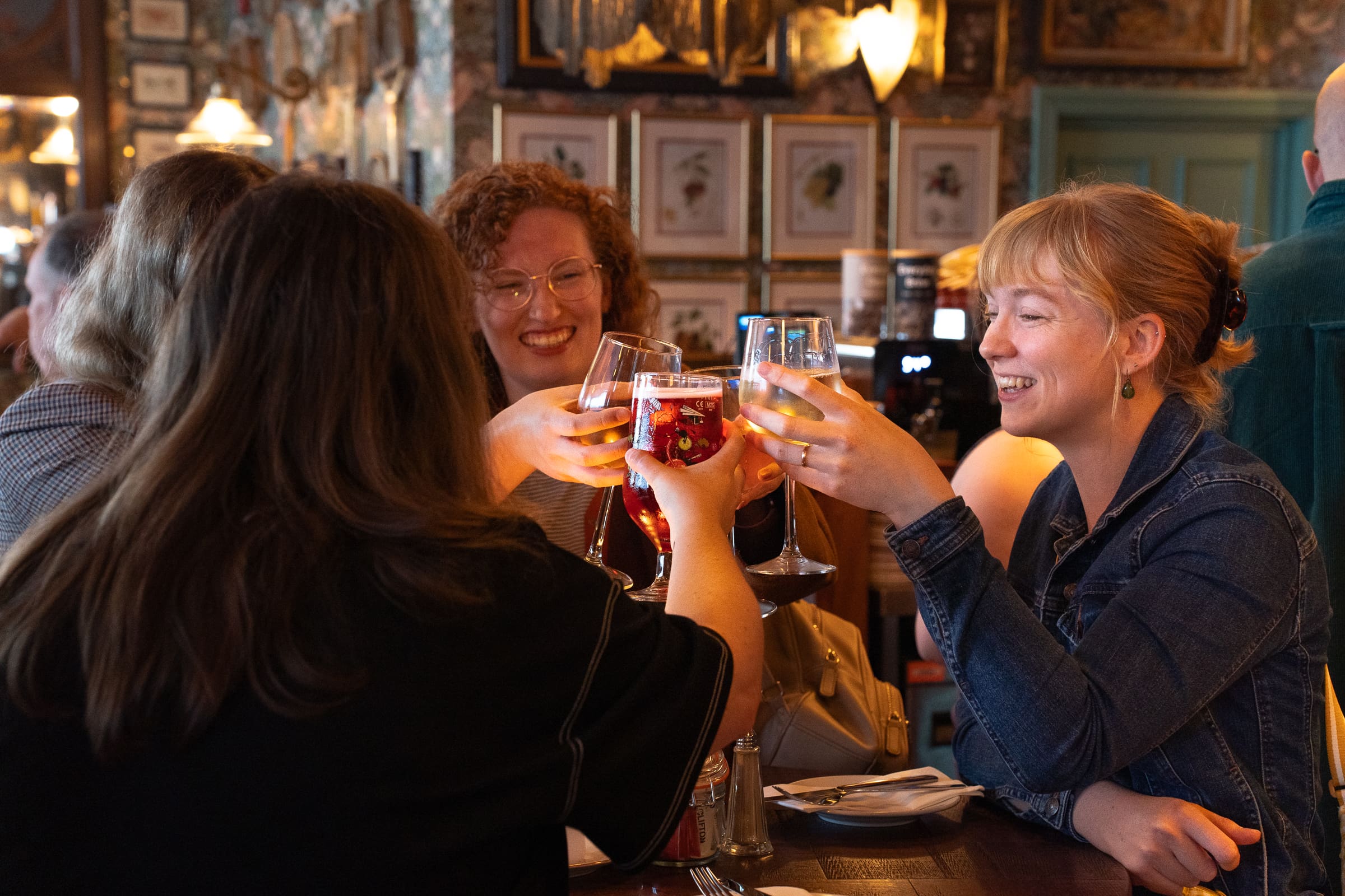 Four friends sit around a table in Salino, a cozy, warmly lit bar, smiling and clinking their drinks together in celebration. The scene feels cheerful and sociable, with artwork and mirrors decorating the walls.