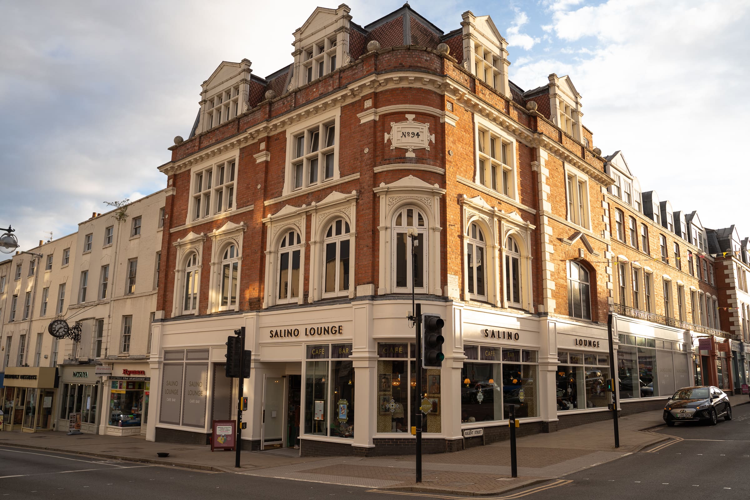 A large, ornate red-brick building with white trim stands on a street corner. The building houses "Salino Lounge" on the ground floor, and sunlight highlights its architectural details. The street is quiet with a few shops nearby.