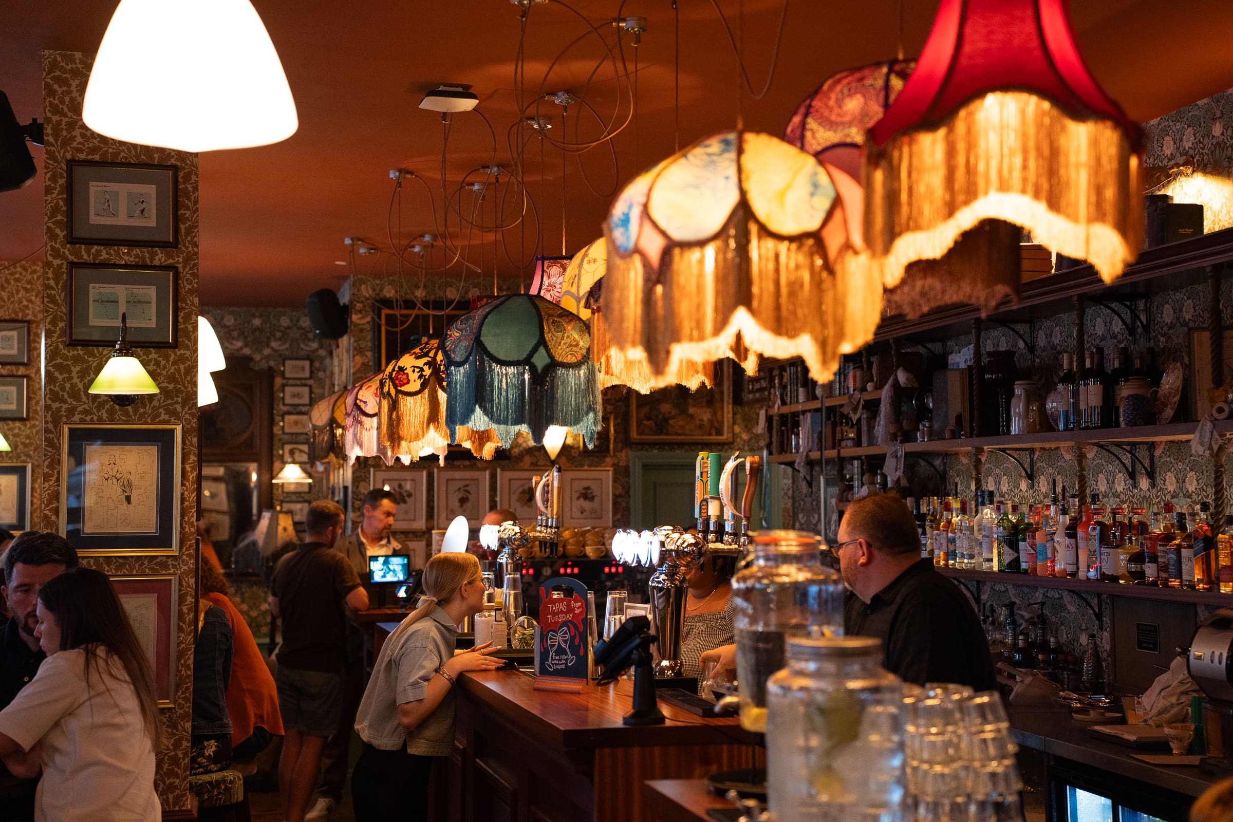 A cozy bar, Salino, with vintage-style lampshades hanging above the wooden counter, people chatting and working behind the bar, and shelves filled with bottles in a warmly lit, patterned interior.