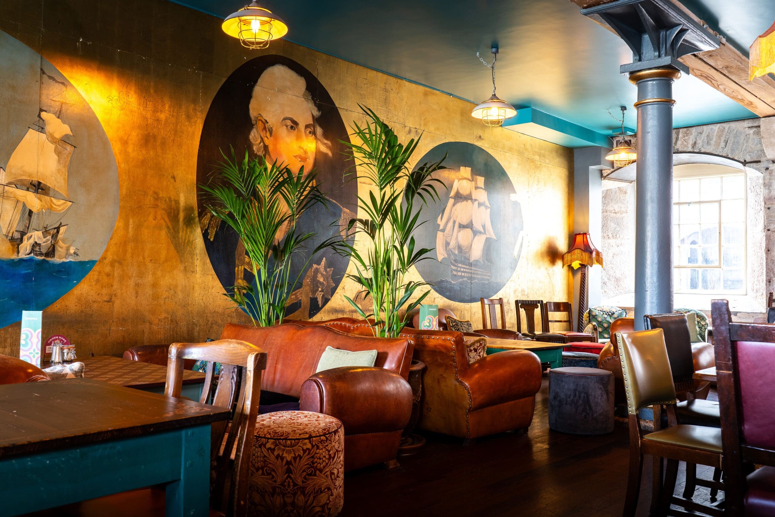 A cozy lounge with leather sofas, wooden tables, and chairs, featuring large nautical-themed murals of ships and Seco, a historical figure, on the golden walls, with indoor plants and soft lighting.