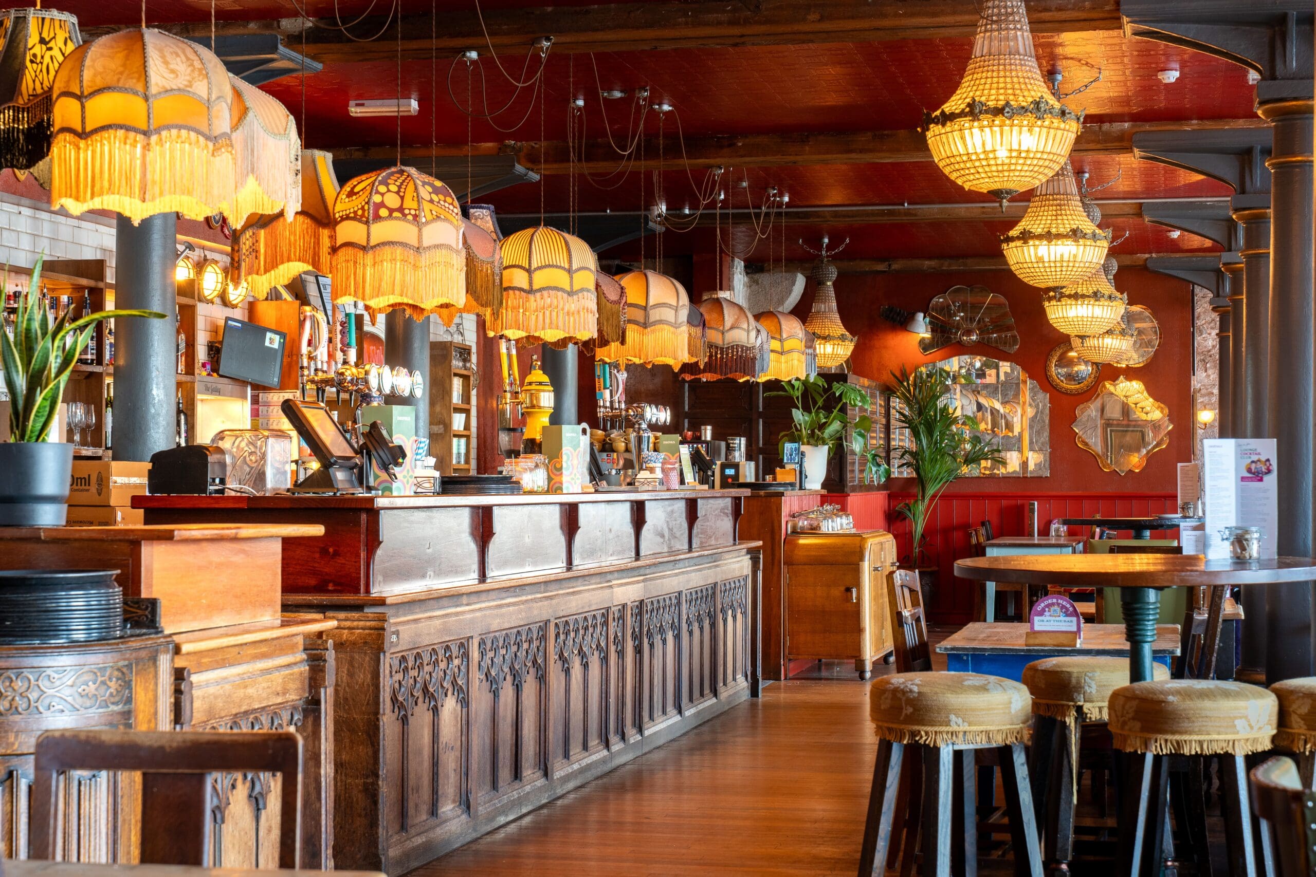 A cozy bar interior features ornate wooden counters, vintage fringed lampshades, high stools, potted plants, and warm lighting—perfect for sipping a classic Seco and enjoying the welcoming, nostalgic atmosphere.