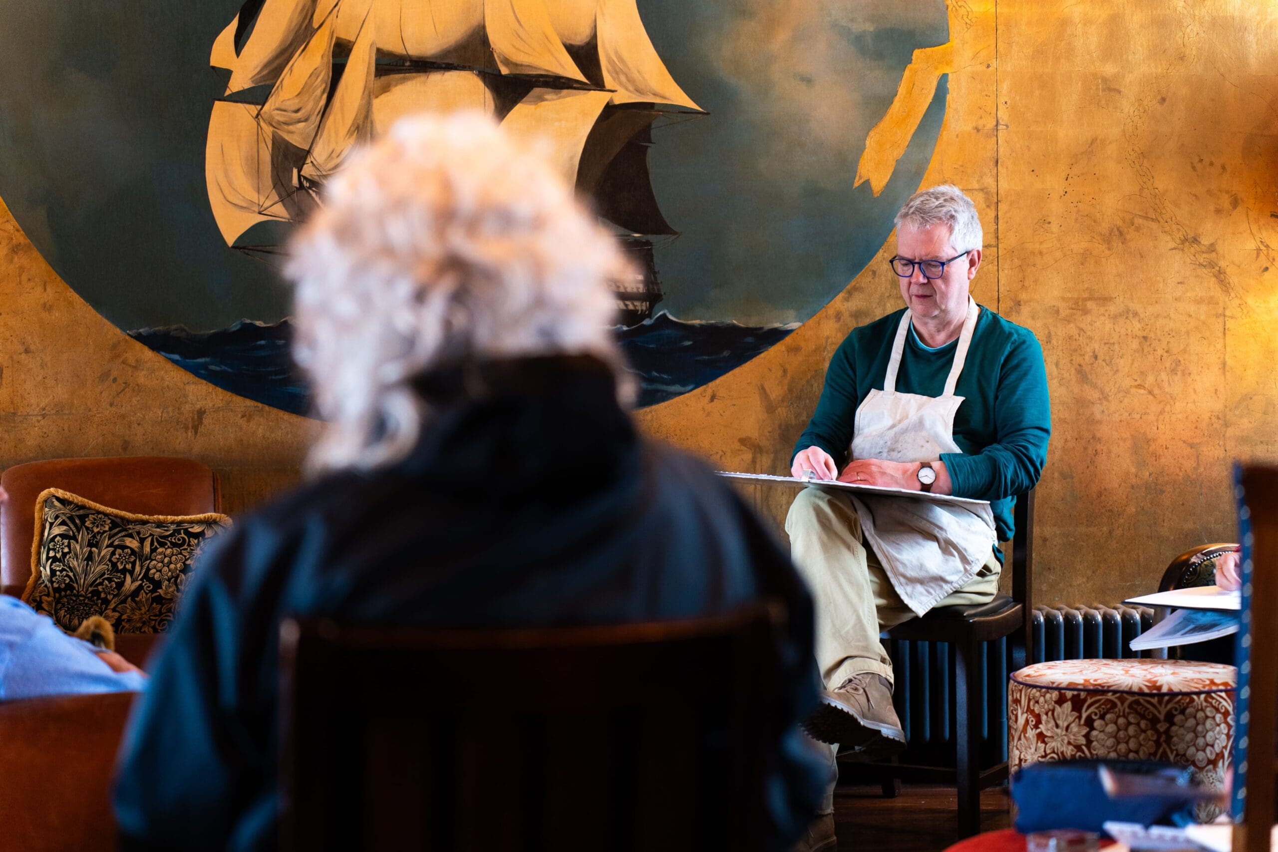 A man in a Seco apron sketches on a pad while seated in a cozy room with gold walls and a large sailing ship mural. Another person with curly white hair sits facing him in the foreground.