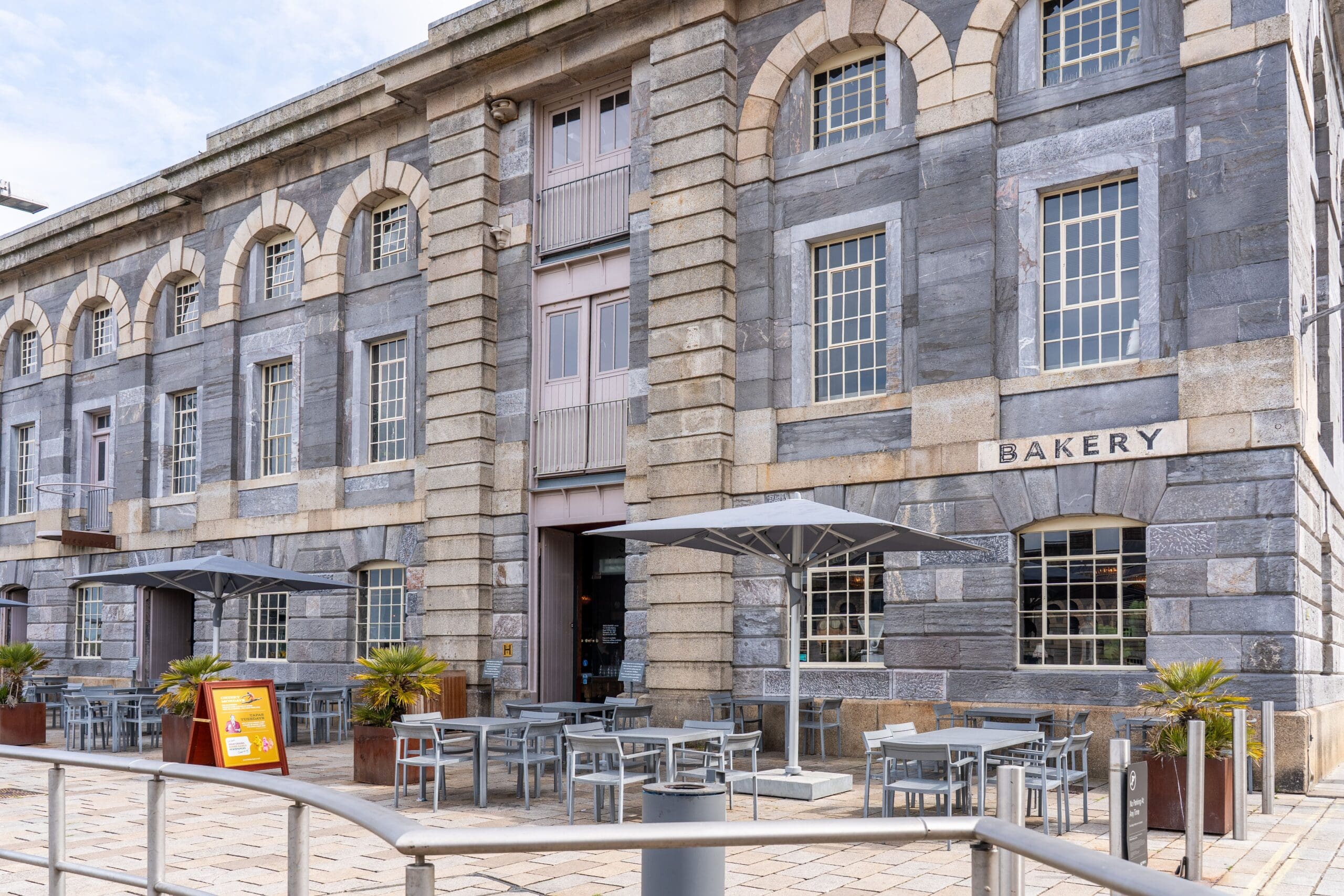 Outdoor seating area with tables, chairs, and umbrellas in front of a gray stone Seco bakery building with large arched windows and a sign that reads "Bakery." A menu board stands near the entrance.