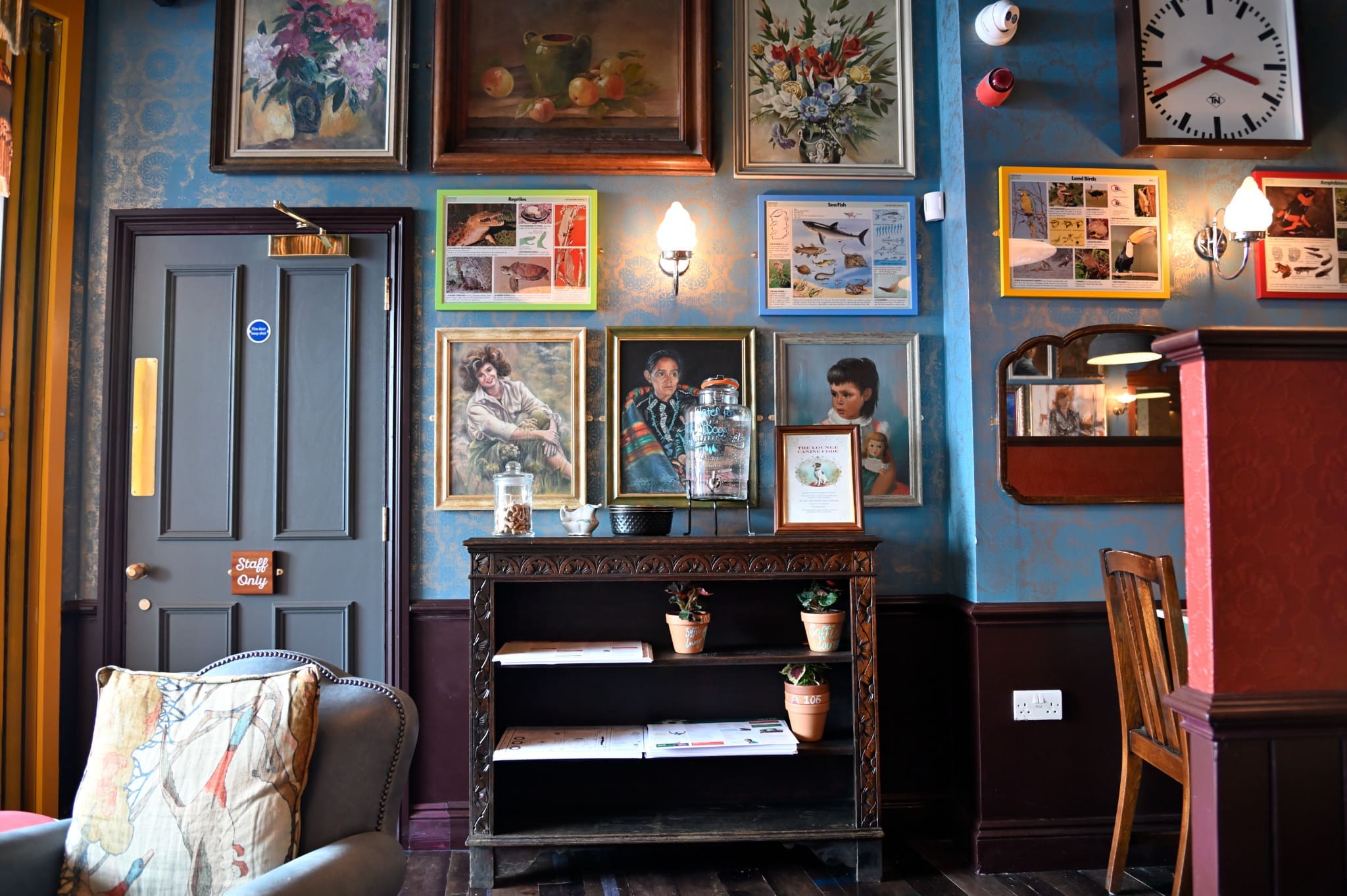 A cozy, vintage-style room with blue patterned wallpaper, framed art and photos, a small Soffio bookshelf adorned with potted plants, a grey door, and a large clock above a wooden chair.