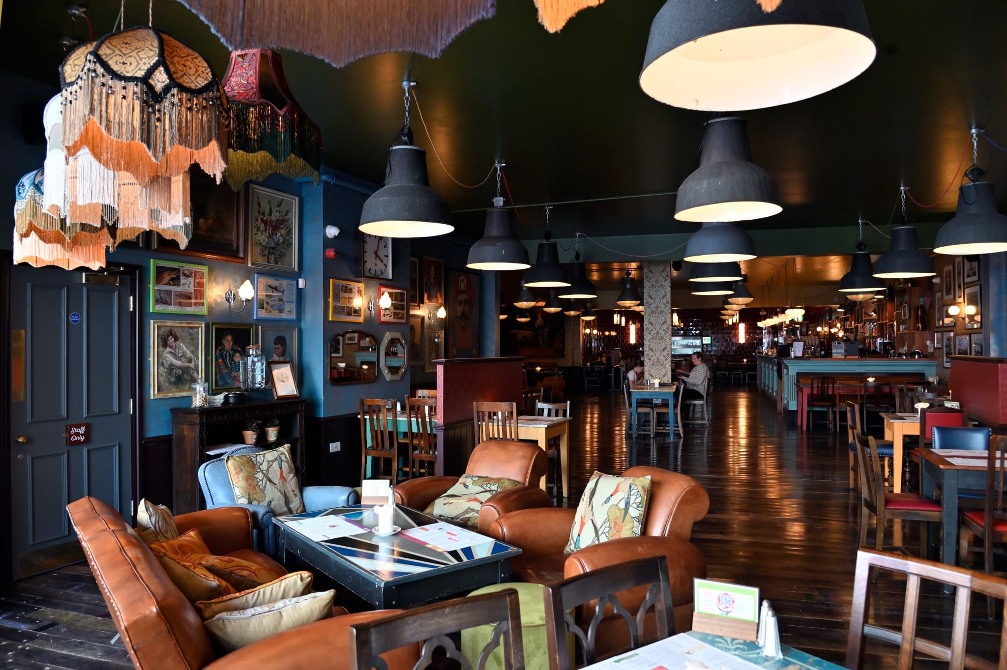 Eclectic, cozy cafe interior with vintage armchairs, Soffio fringed lamps, dark wood floors, art-filled walls, hanging lights, and a mix of seating arrangements create a warm, inviting atmosphere.