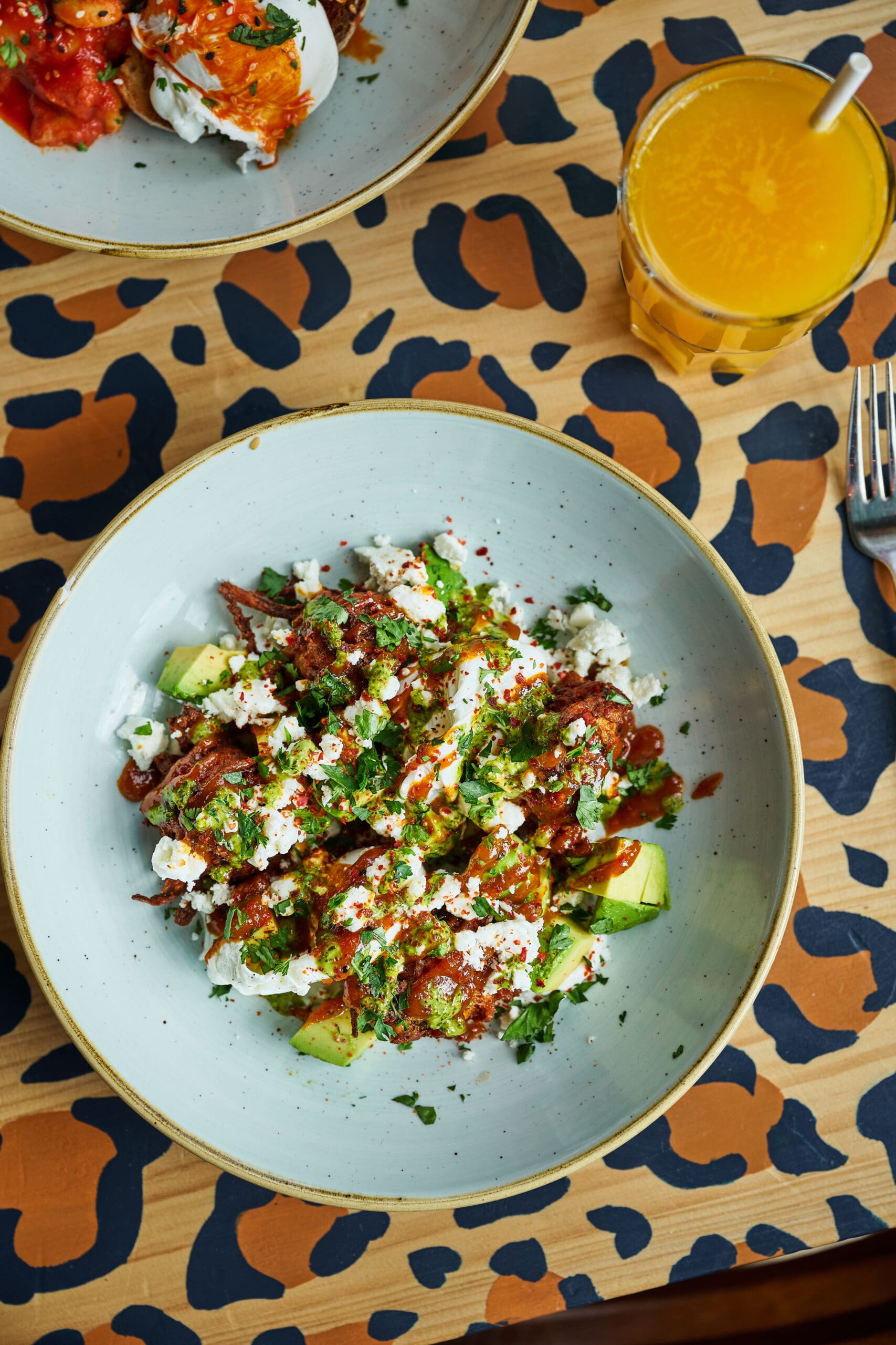 A bowl of salad with avocado, herbs, and sauce sits on a table with a bold, leopard-print pattern beside a glass of orange juice. A fork rests nearby.