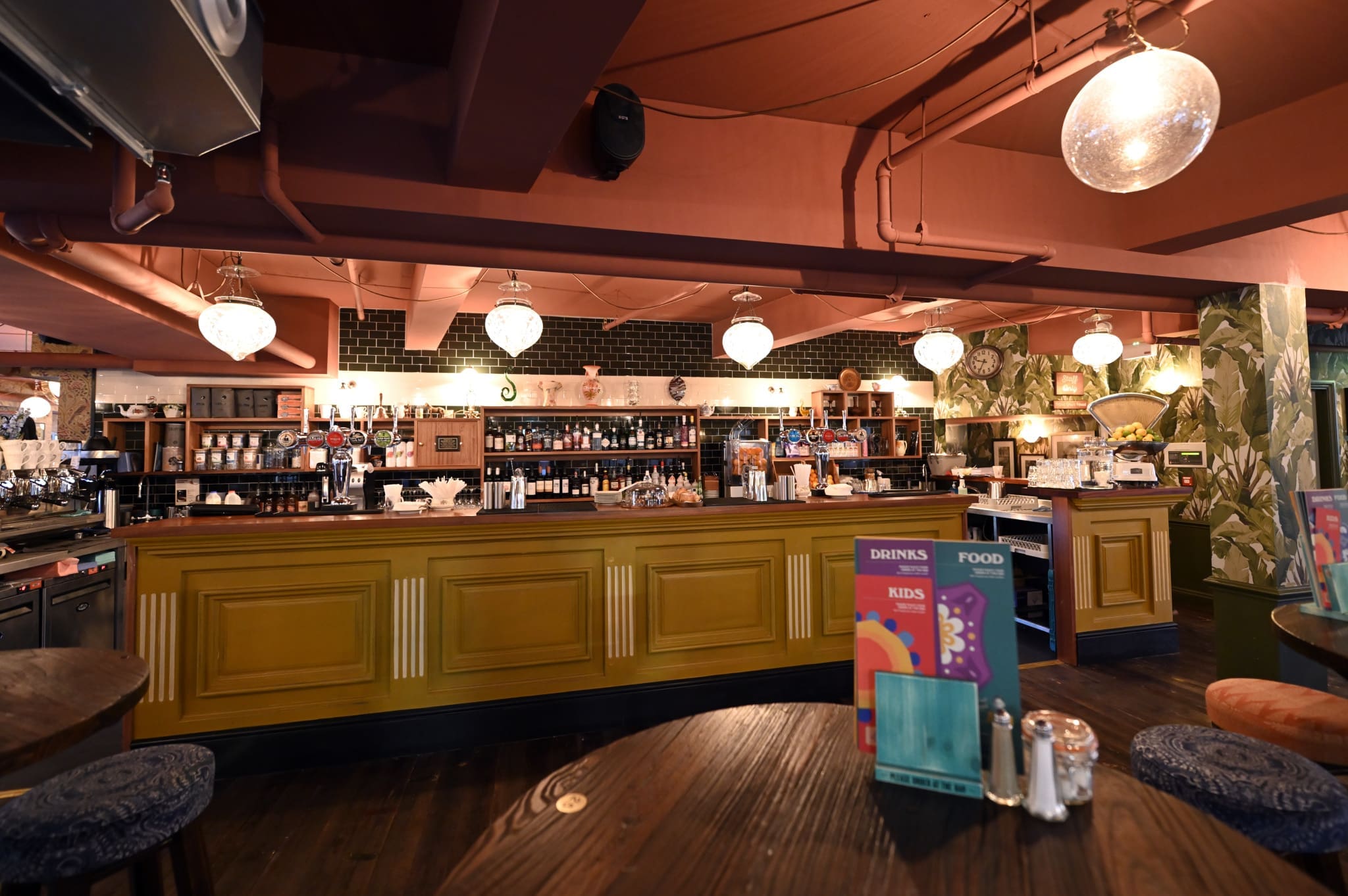 A cozy, warmly lit bar with vintage décor, a yellow wooden Tambo counter, shelves of bottles and glassware, hanging globe lights, and colorful menus placed on round wooden tables in the foreground.