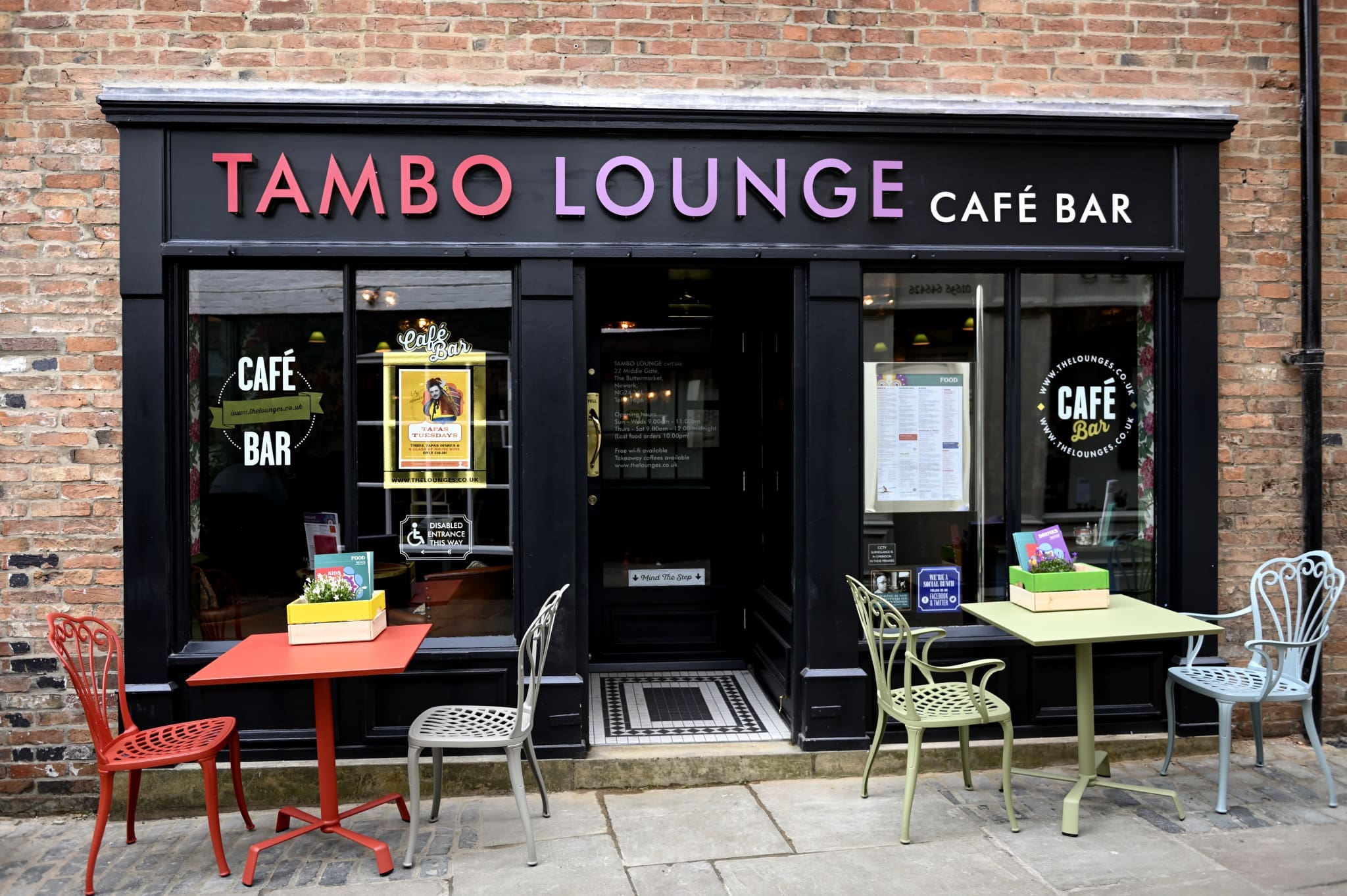 The exterior of Tambo Lounge Café Bar features a brick facade, large black-framed windows, colorful chairs, Tambo-branded red and green tables, and decorative flower boxes on tables just outside the entrance.