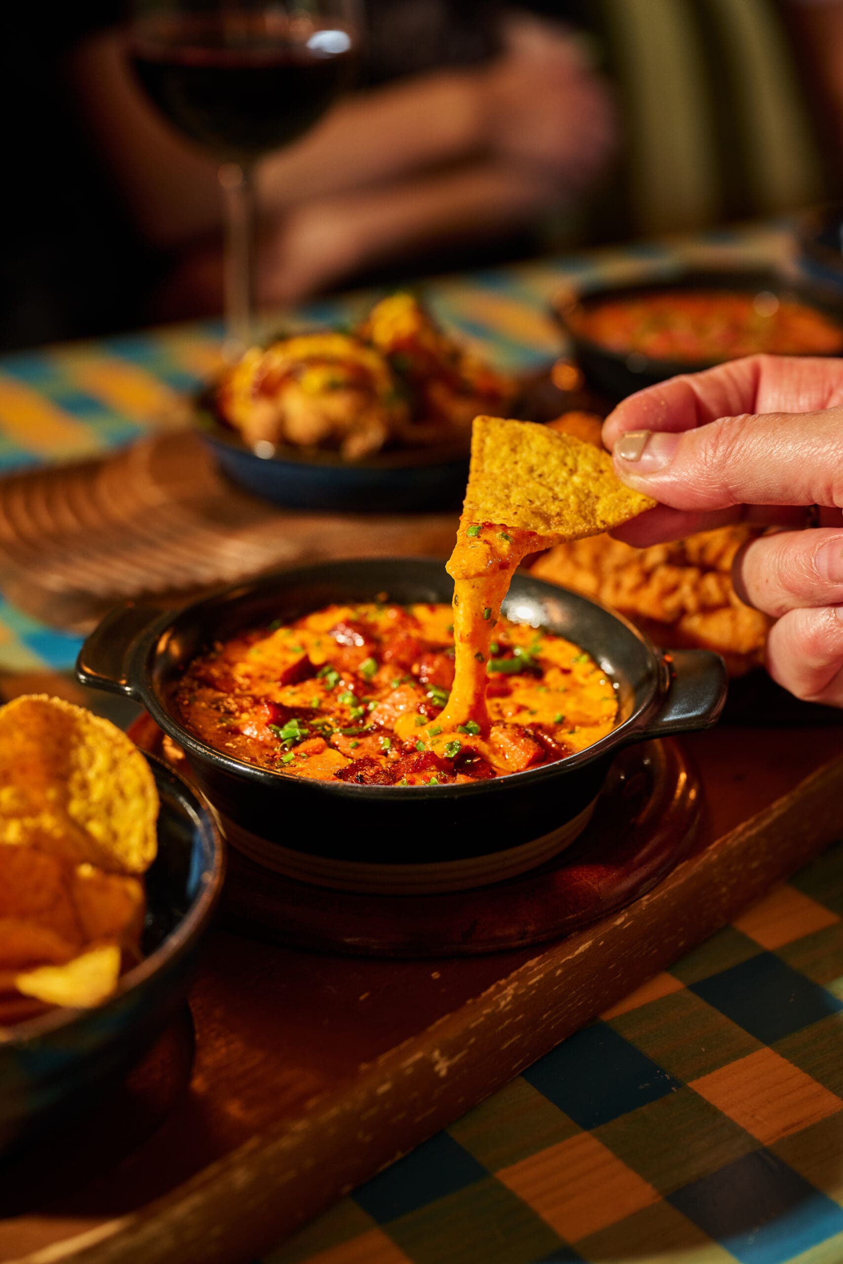 A hand dips a tortilla chip into a bowl of melted, cheesy dip with herbs and tomato on top. Other dishes and a glass of red wine are blurred in the background on a checkered table.