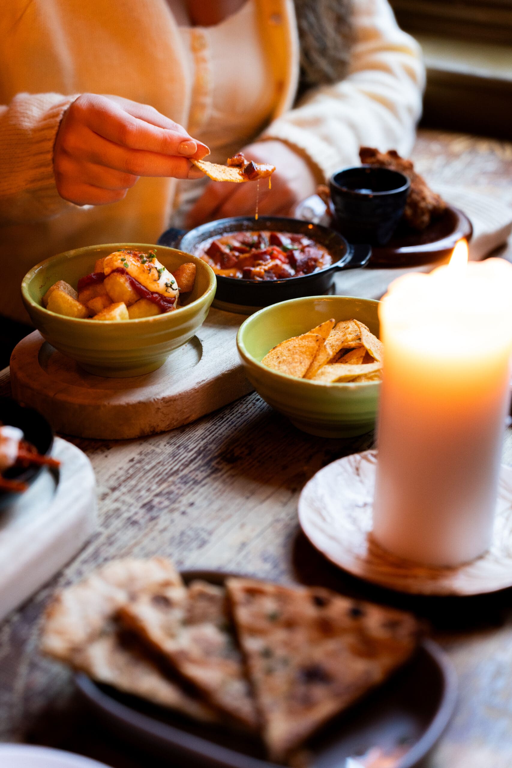 A person lounges at a wooden table, dipping a chip into a bowl of salsa amid various dishes—sliced bread, chips, potatoes—and the warm glow of a lit candle.