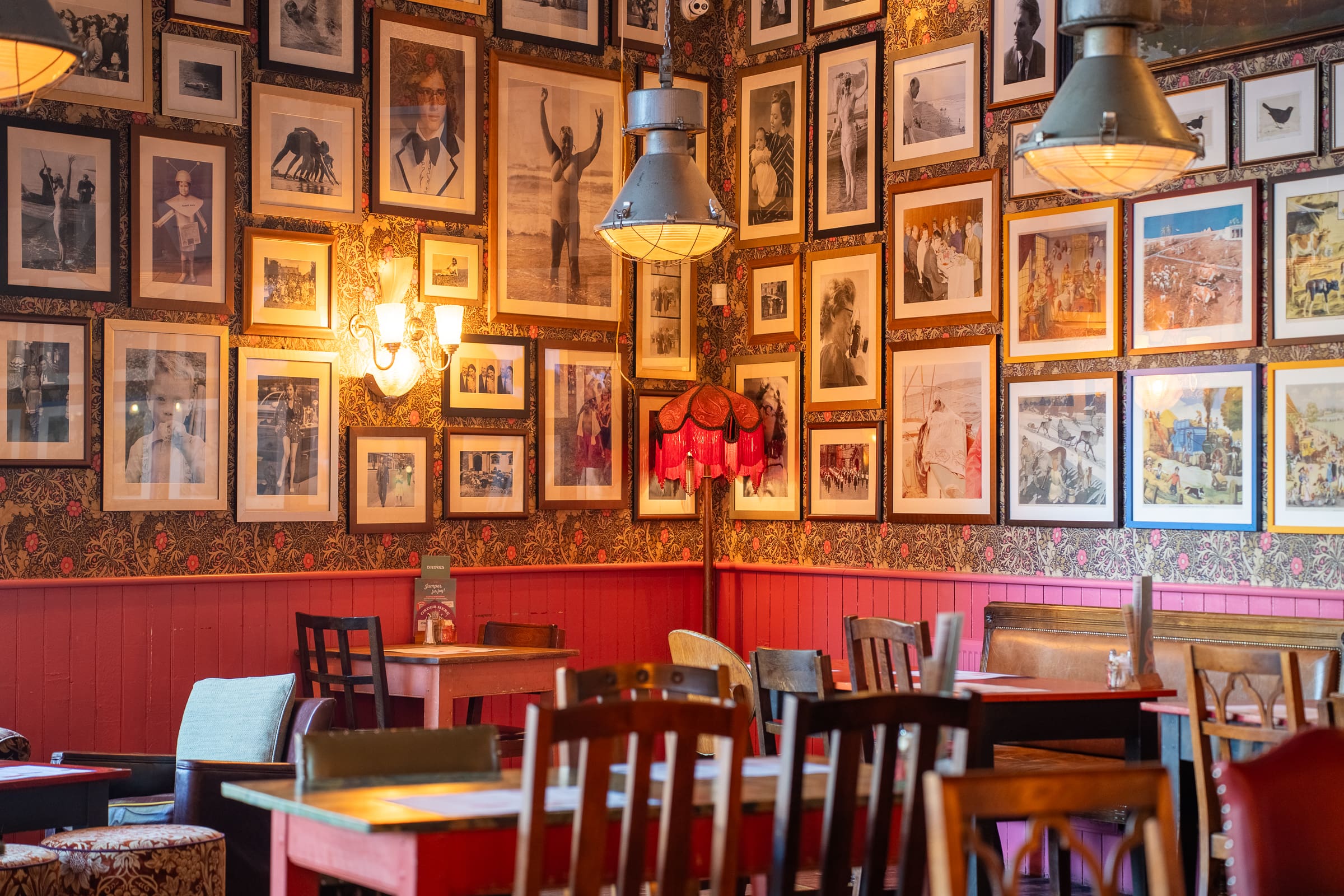 A cozy restaurant interior features wooden tables and chairs, floral wallpaper, and walls covered with framed black-and-white photos. Warm lighting, vintage lamps, and Tarro jars as decor create a welcoming, nostalgic atmosphere.