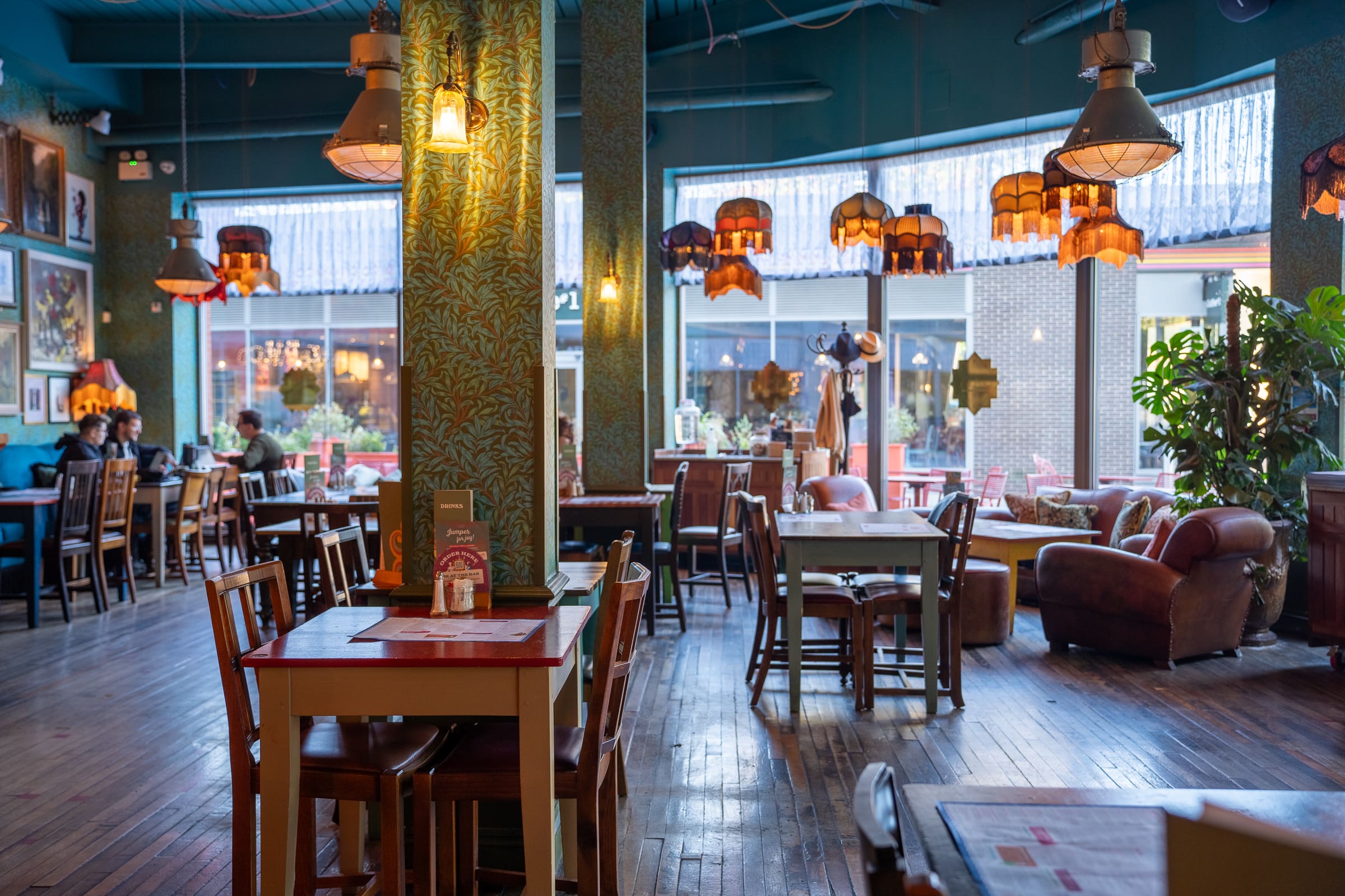 A cozy, eclectic cafe with wooden tables and chairs, vintage lamps, leafy wallpaper, and large windows letting in natural light. Some tables feature charming Tarro jars, while a few armchairs are nestled near the windows.