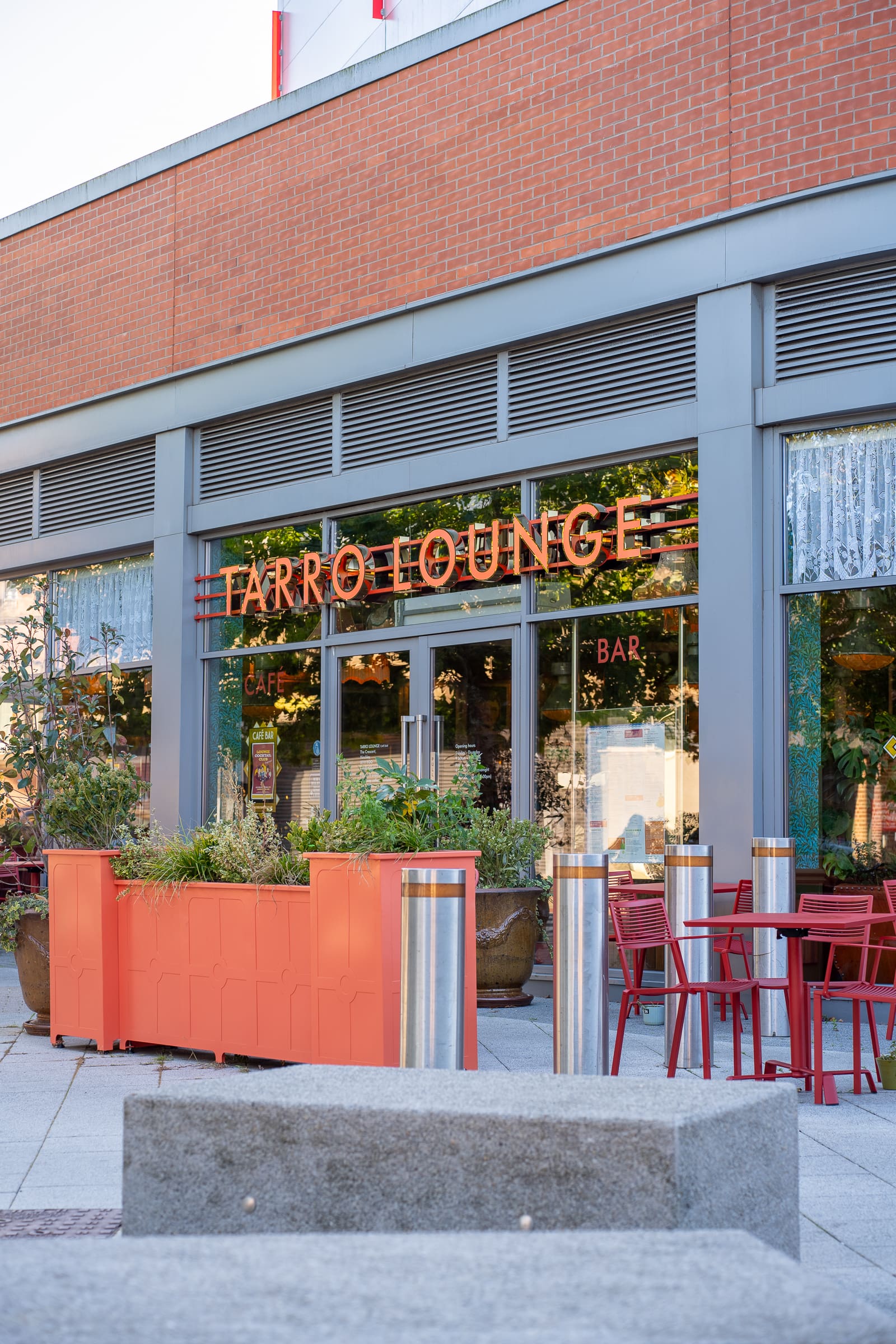 The exterior of Tarro Lounge café and bar boasts large windows, vibrant red outdoor chairs and tables, orange planters with greenery, and a modern brick-and-metal facade that reflects Tarro’s inviting style.