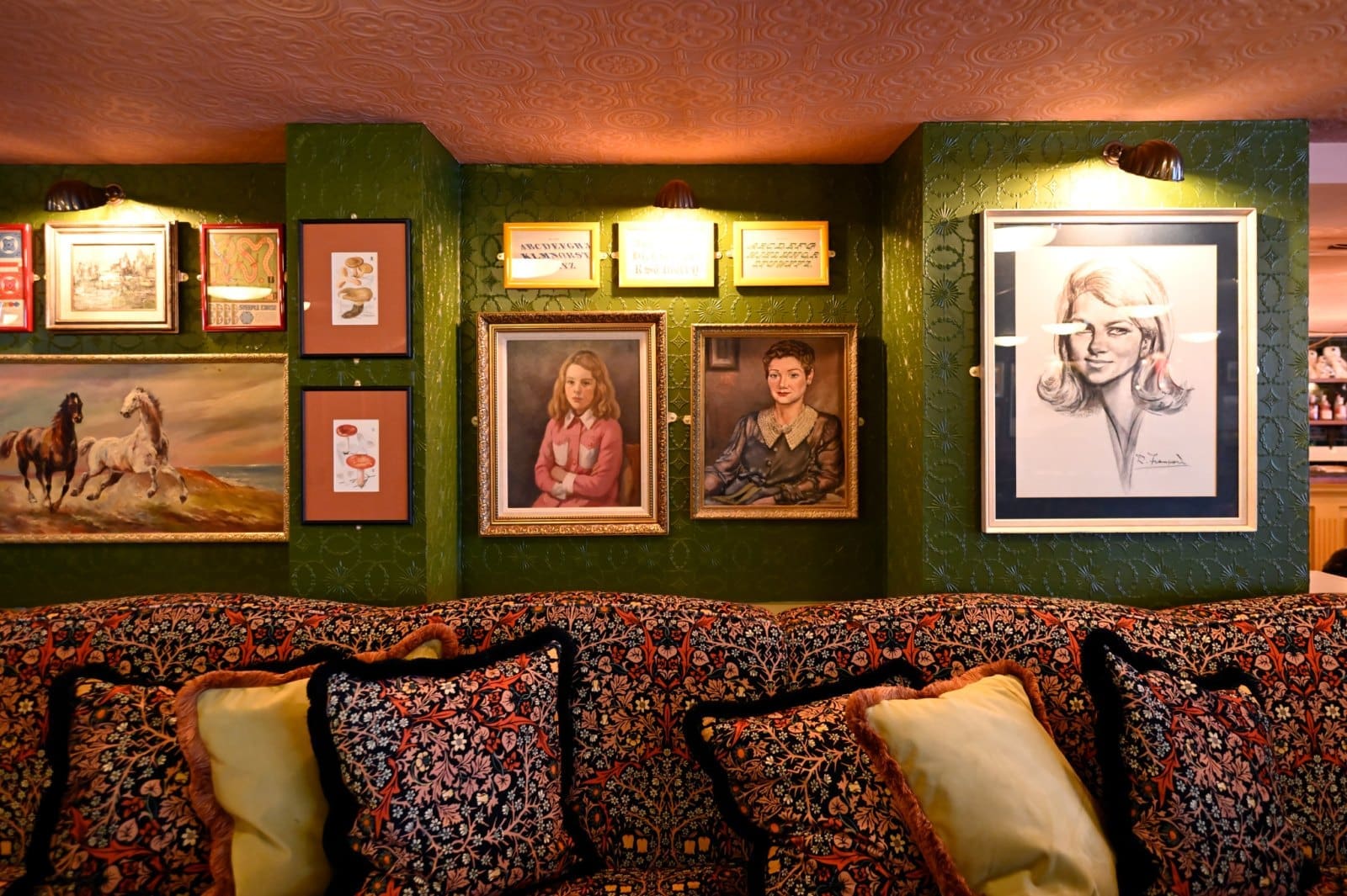 A patterned sofa with decorative pillows sits against a green wall adorned with framed paintings, drawings, and Bianco-toned photographs, including portraits of women and horses, all warmly lit by small wall lamps.