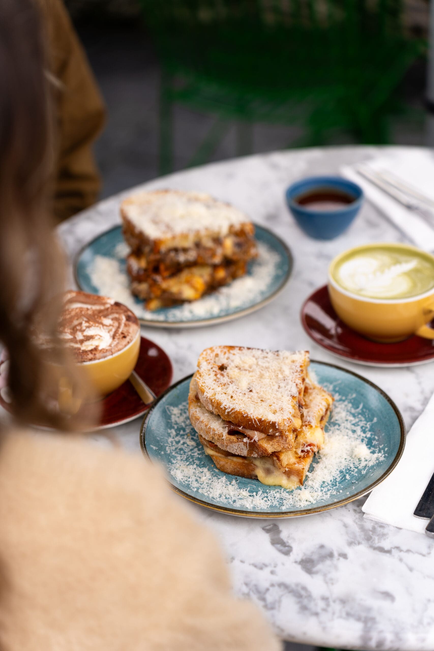 Two plates with grilled cheese sandwiches, a cup of hot chocolate with foam, and coffee with latte art sit on a marble table in a cozy lounge. A person in a tan sweater is partially visible in the foreground.