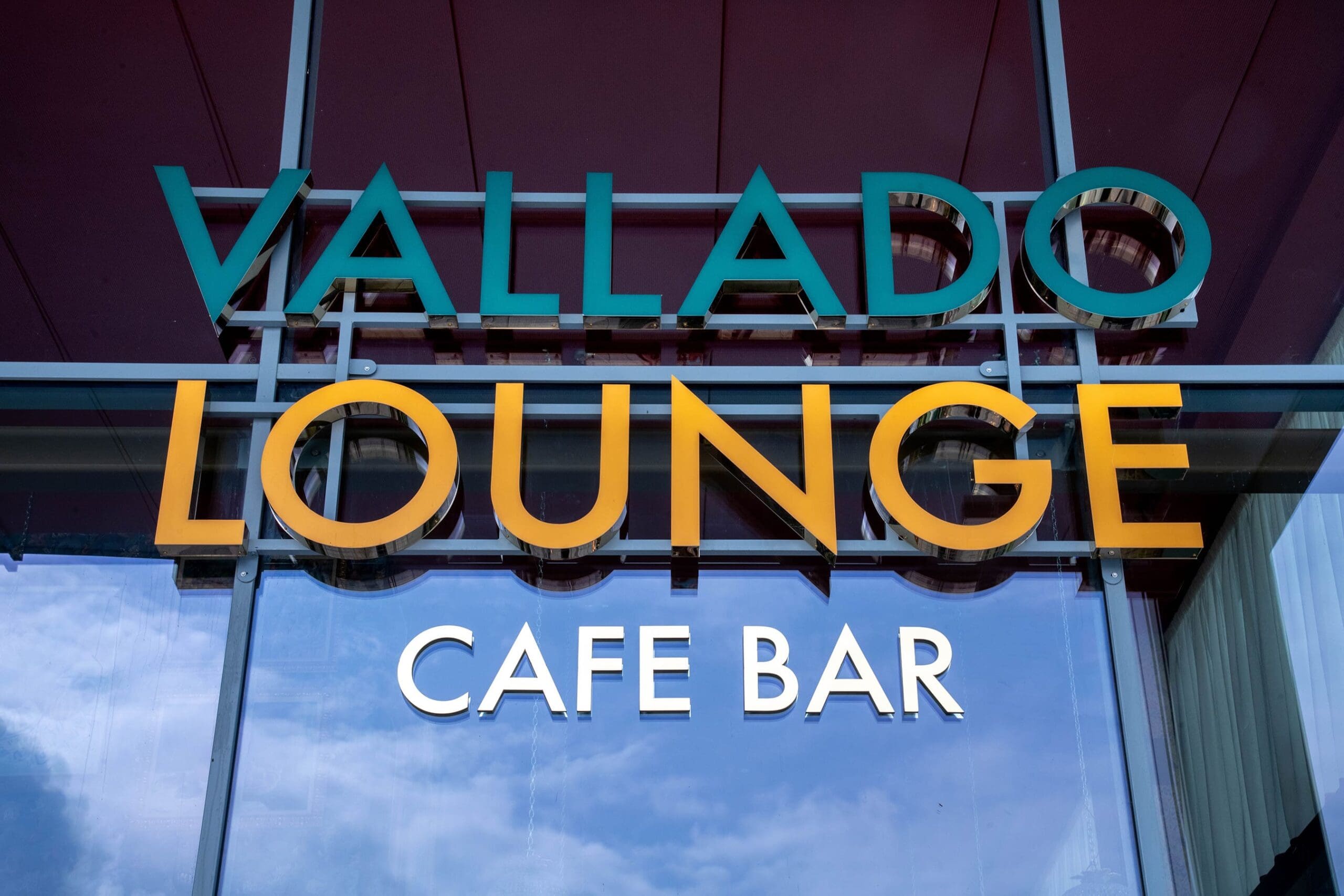 Glass storefront with bold green and yellow letters spelling “VALLADO LOUNGE” and white letters below for “CAFE BAR,” reflected against a blue sky with clouds, adds a touch of vitello charm to the inviting atmosphere.