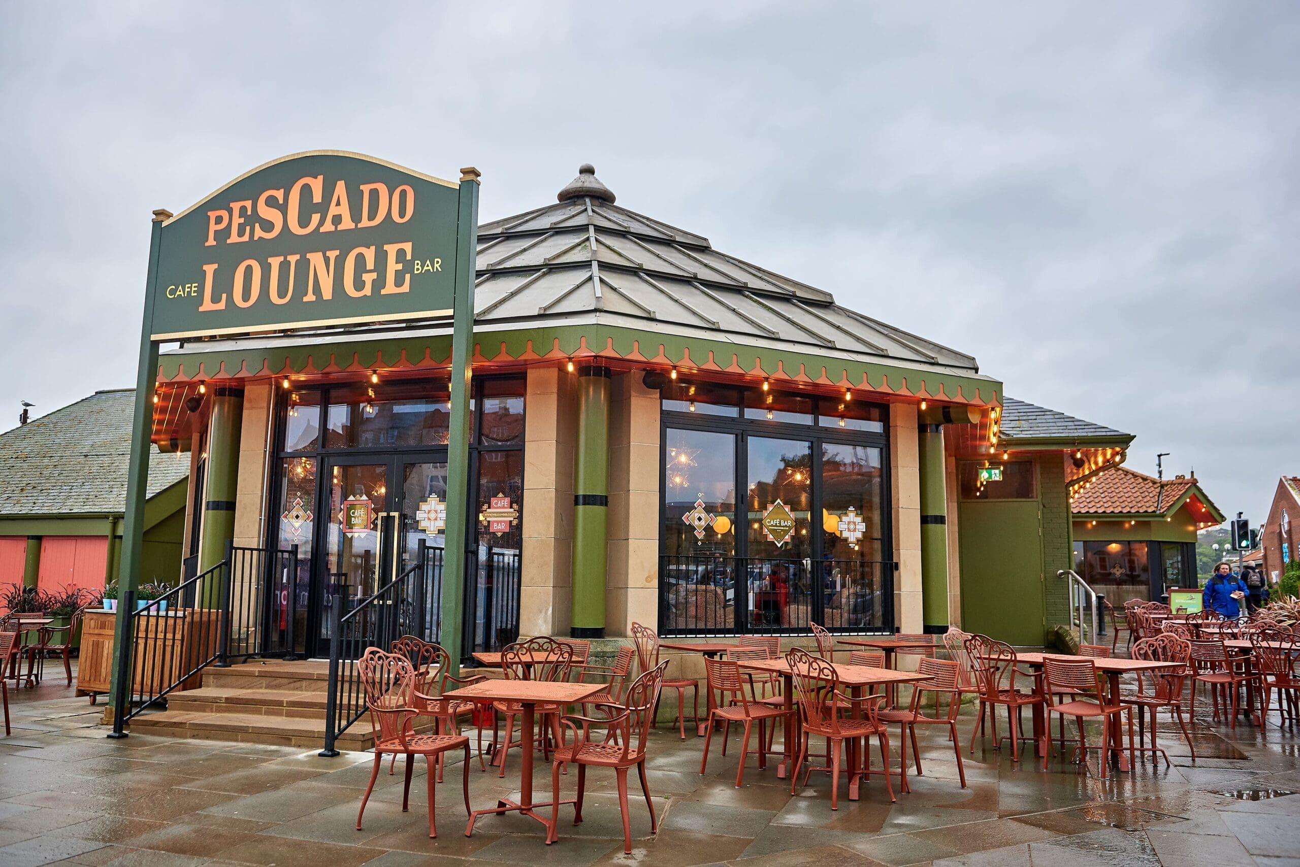 Outdoor view of Pescado Lounge café and bar with large windows, a round building, green pescado sign, and empty red tables and chairs on the wet pavement under a cloudy sky.