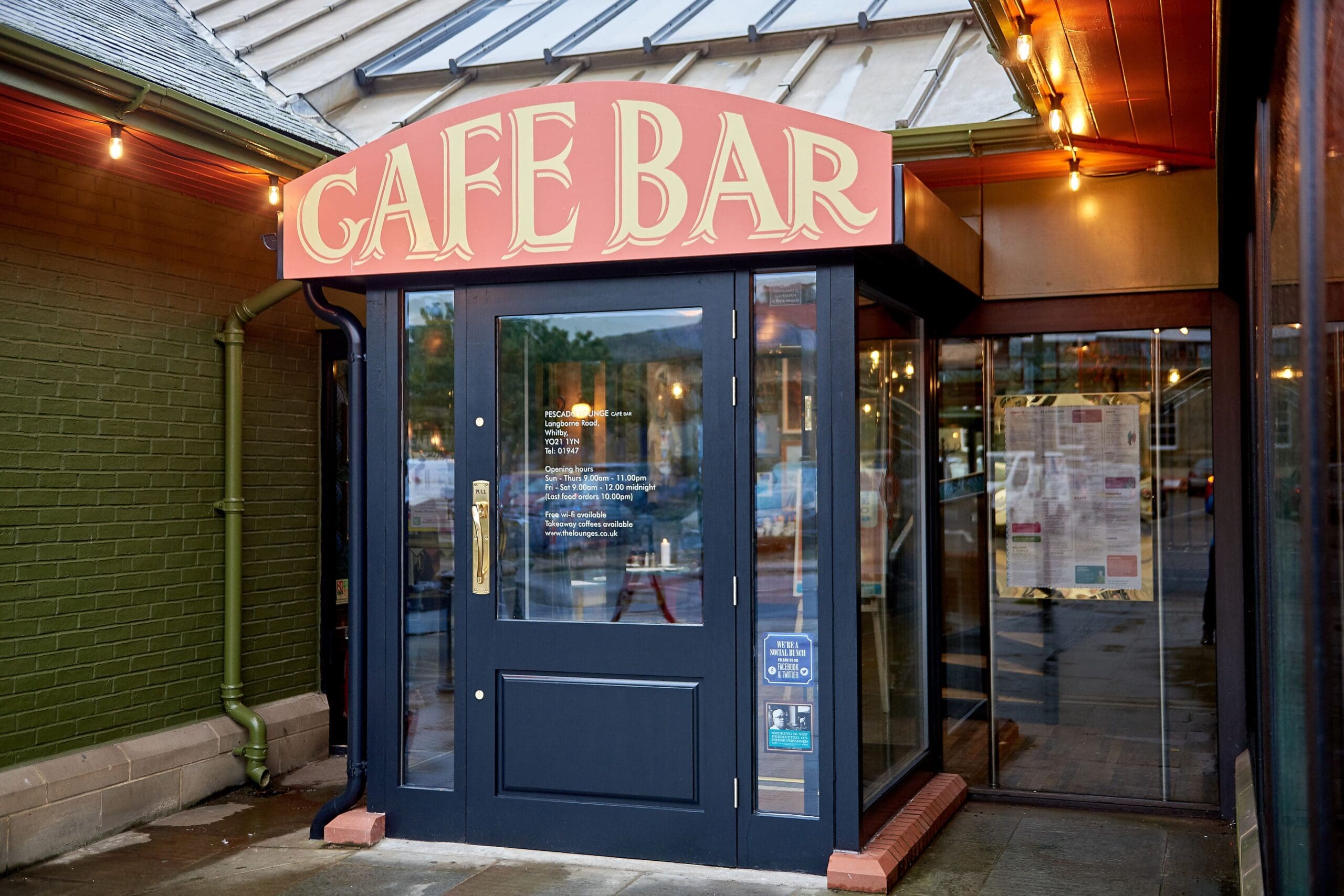 Entrance to a café bar with a red and yellow sign above glass doors; lights hang overhead, reflections in the windows hint at the café’s specialty—perhaps even fresh pescado on the menu.