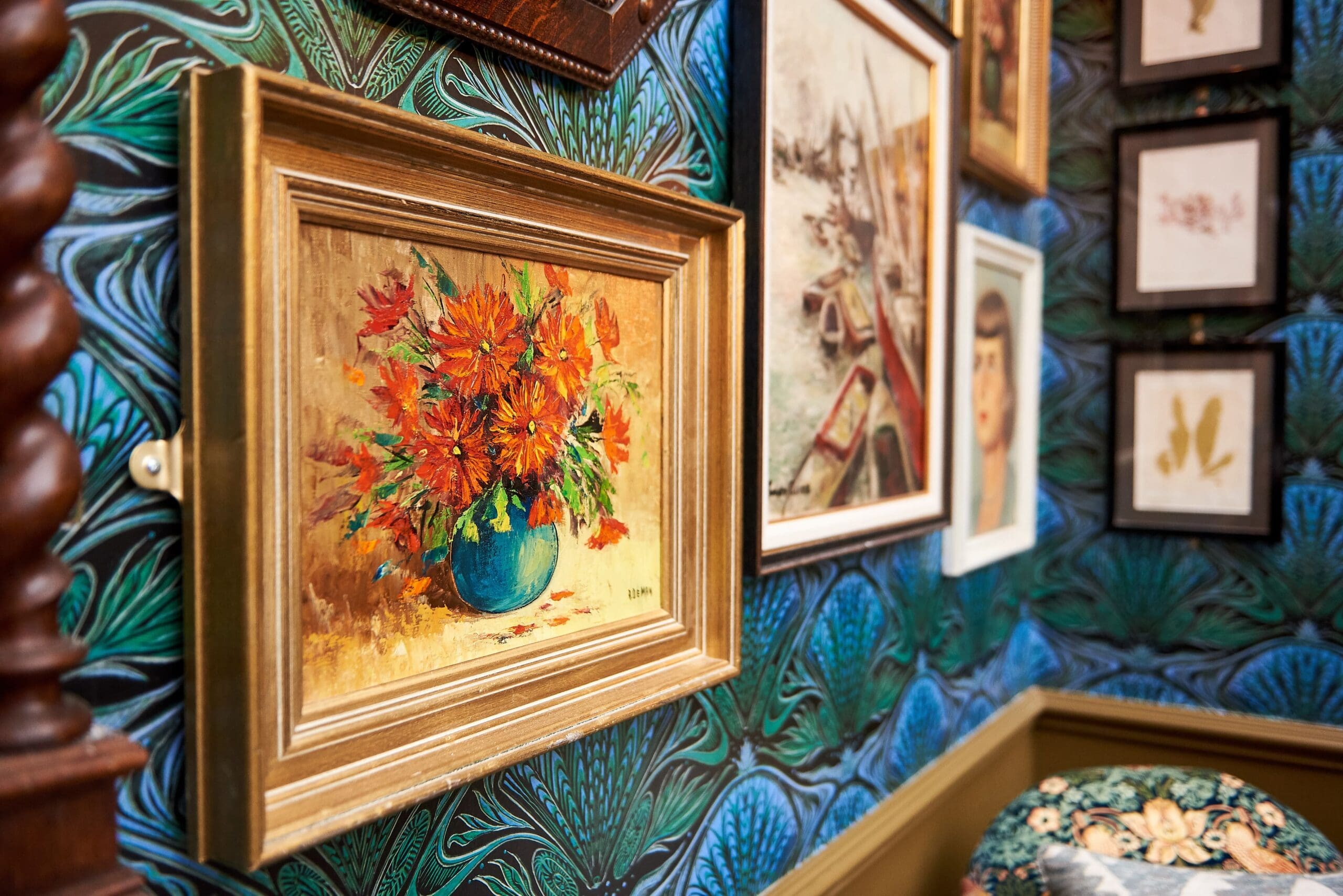 A close-up of a gallery wall with framed paintings, including a vibrant floral still life and a delicate pescado illustration in gold frames, set against blue patterned wallpaper with green leafy designs.