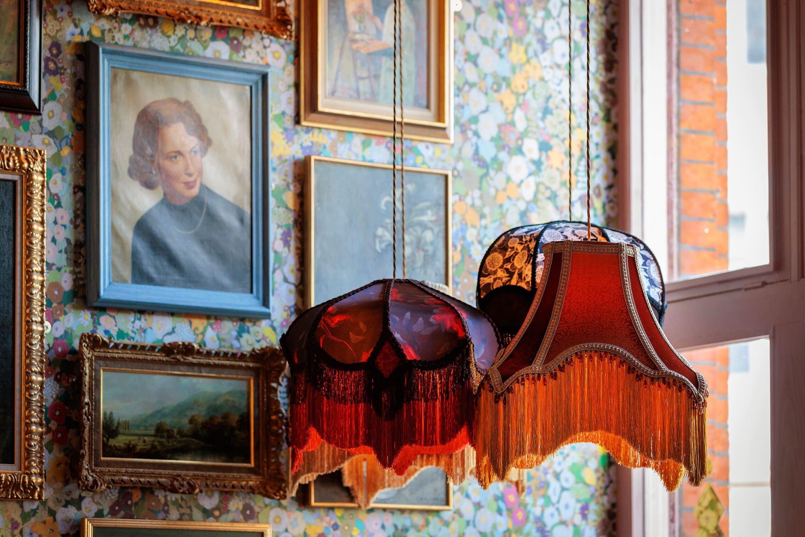 Two vintage lamps with fringe hang in front of a floral wallpapered wall, decorated with framed paintings—including a striking portrait of a woman, several landscapes, and a subtle leo motif—near a window bathed in natural light.