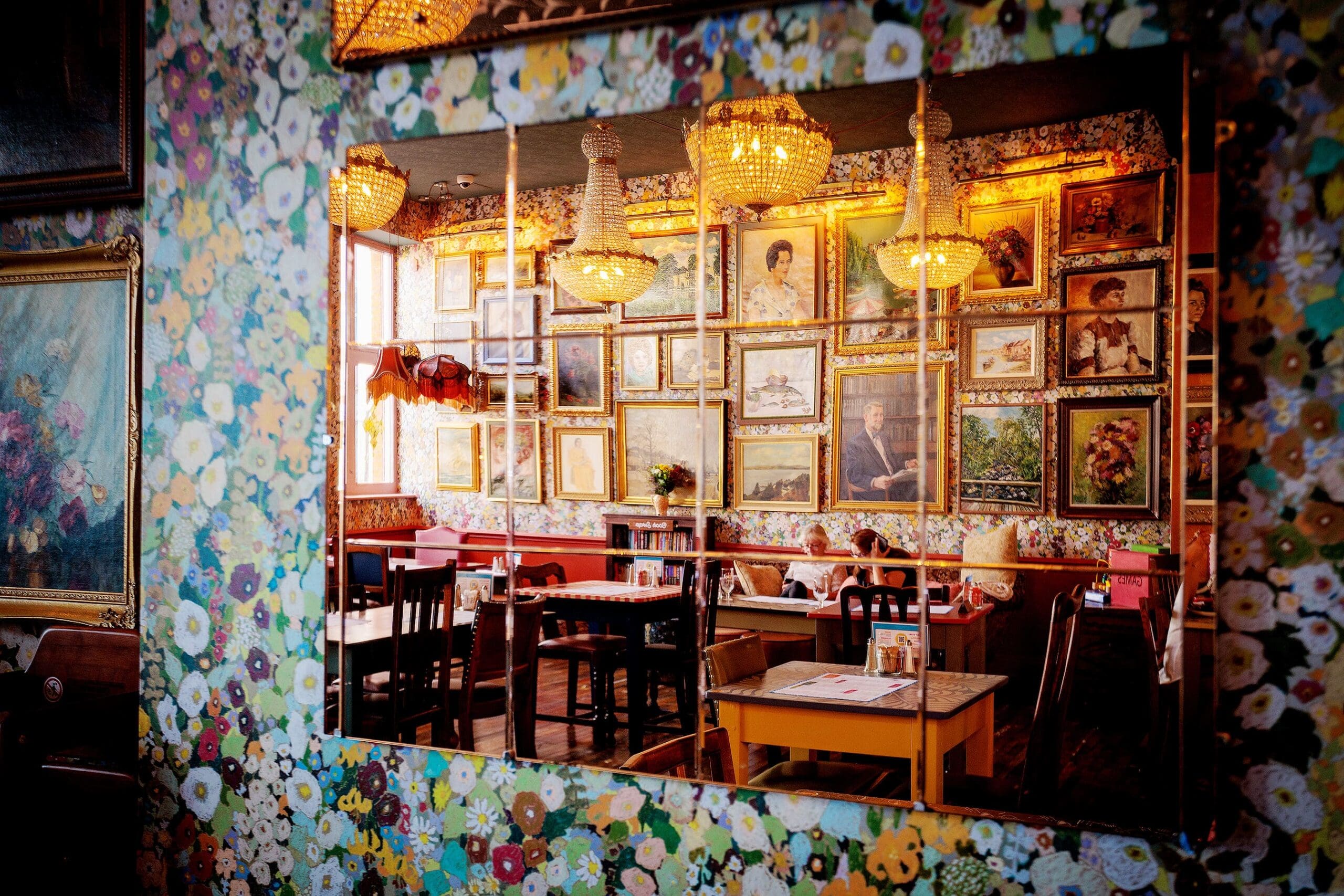 A large wall mirror reflects a cozy, eclectic cafe decorated with floral wallpaper, chandeliers, and framed art—including a striking Leo motif. Tables and chairs fill the space, creating a vibrant, inviting atmosphere.