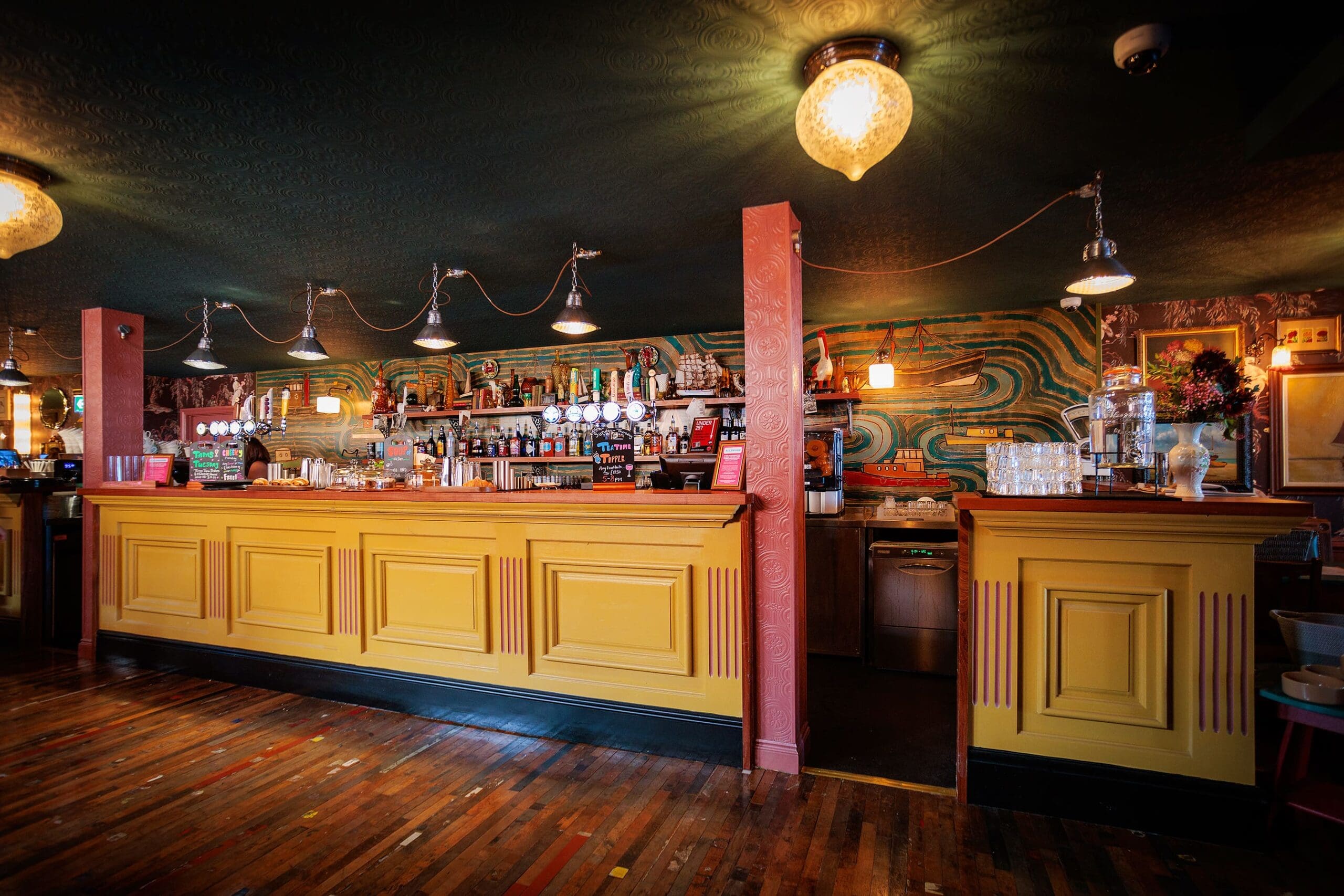 A vibrant Leo-inspired bar with a yellow wooden counter, colorful wallpaper, and shelves stocked with bottles. The dark ceiling and decorative hanging lights set the scene, while the rustic wooden floor adds to the inviting atmosphere.