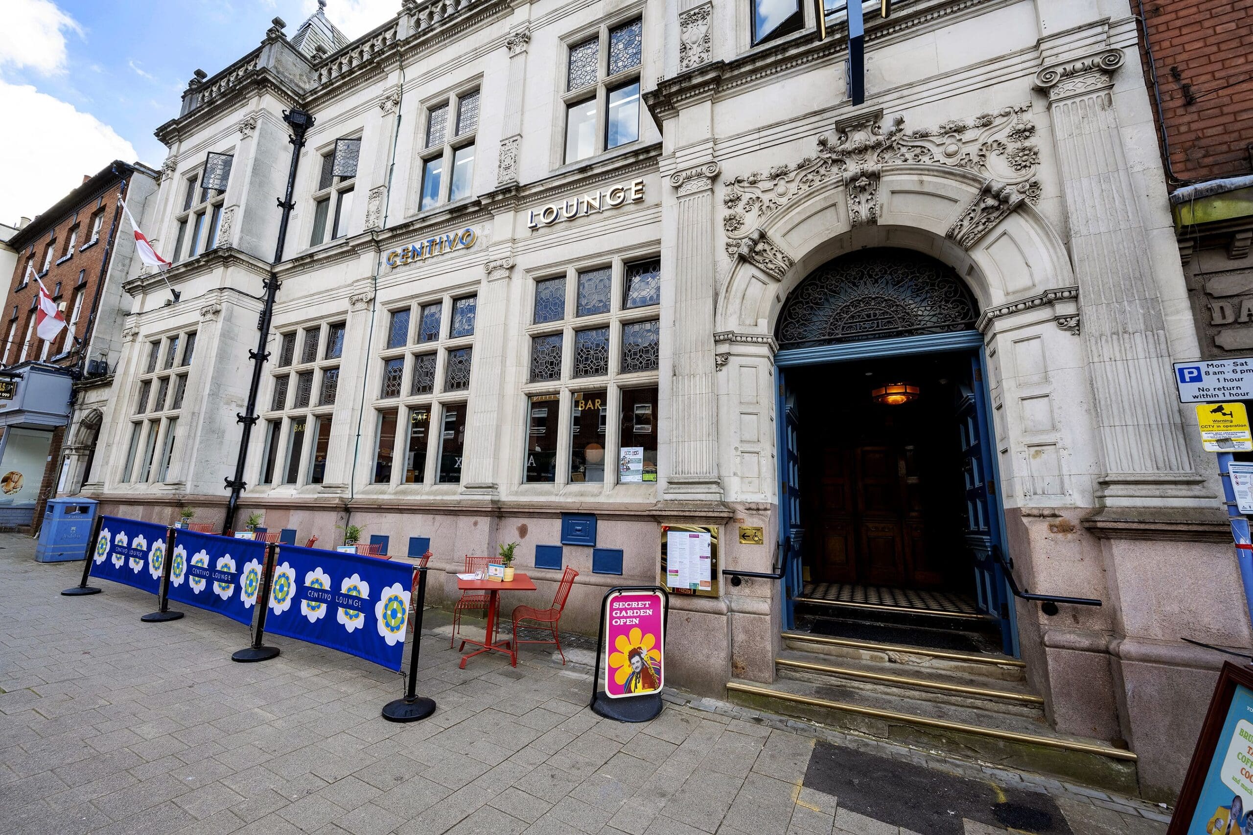 Street view of a grand historic building with "Crafting Lounge" signage and outdoor tables; centivo posters and signs are visible on the building and sidewalk, while an open blue door invites entry past decorative barriers.