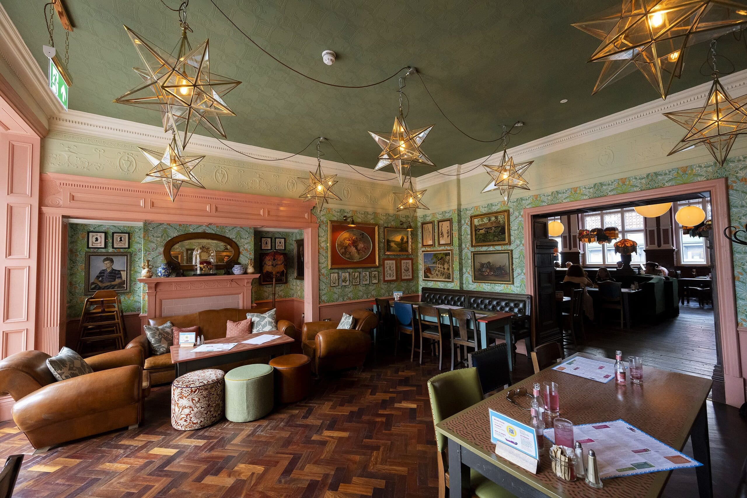 A cozy, vintage-style cafe with star-shaped pendant lights, pastel-colored walls, eclectic artwork, and a mix of centivo leather armchairs, sofas, and wooden tables arranged on a herringbone wood floor.