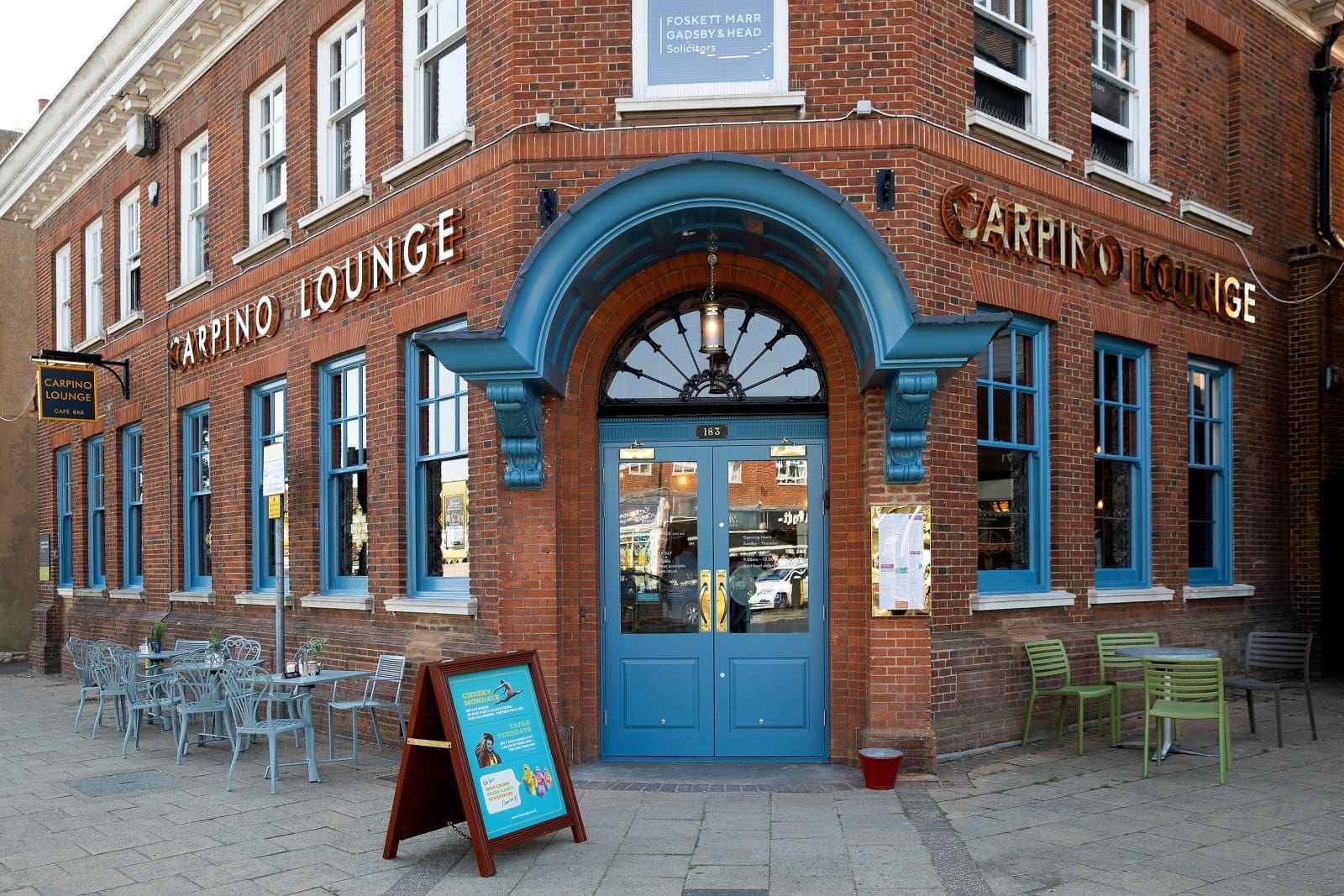 Carpino Lounge is a brick café with blue doors and windows, cozy outdoor tables and chairs, and a sandwich board sign welcoming guests on the pavement in front of the entrance.