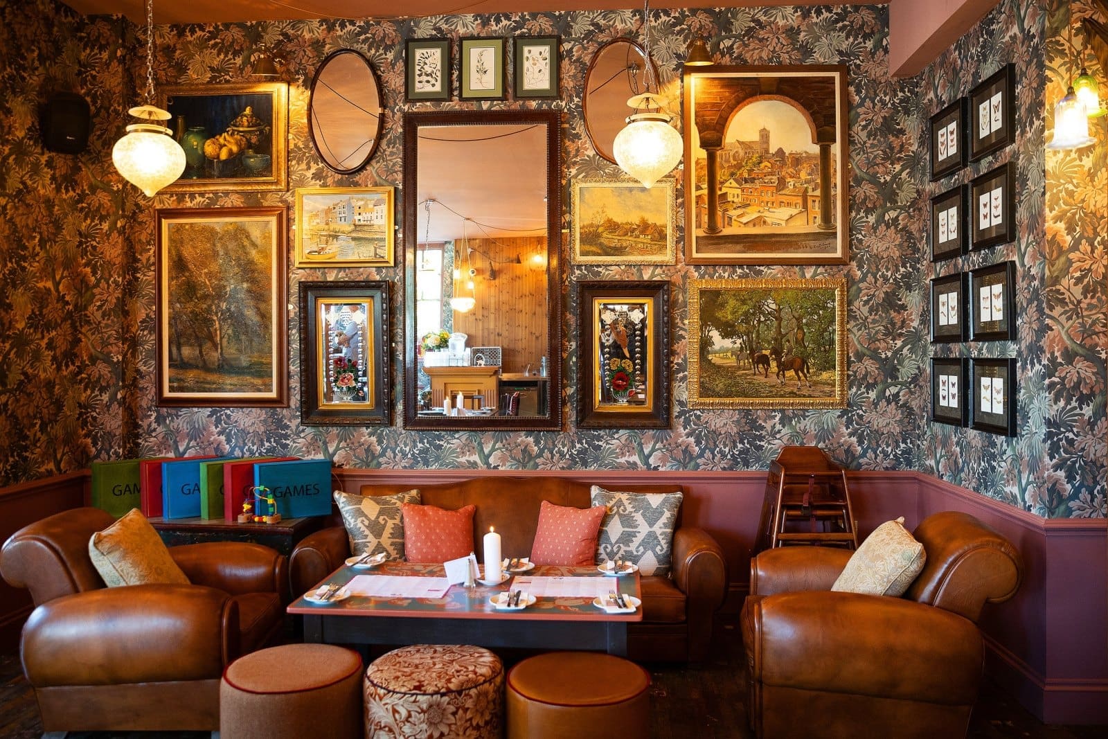 A cozy, vintage-style lounge with floral wallpaper, leather armchairs, framed artwork and mirrors on the wall, a small carpino table set for tea, and soft, warm lighting.