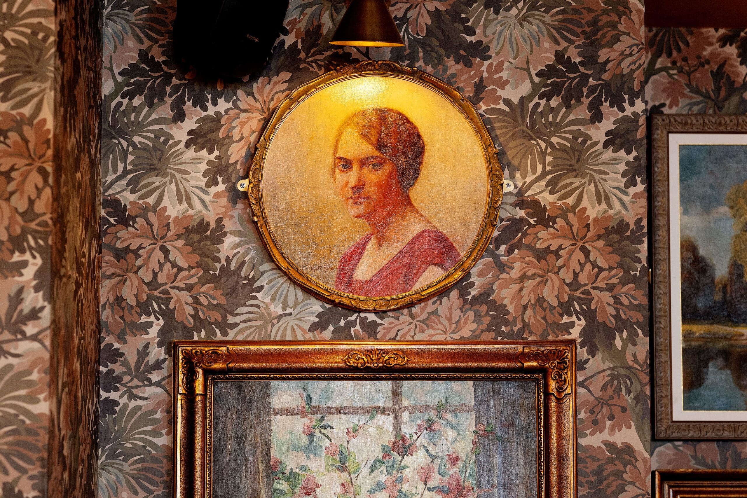 A framed oval portrait of a woman with brown hair hangs on a patterned floral wallpapered wall above another framed painting depicting flowers. Warm light shines on the portrait, creating a cozy carpino-inspired atmosphere.