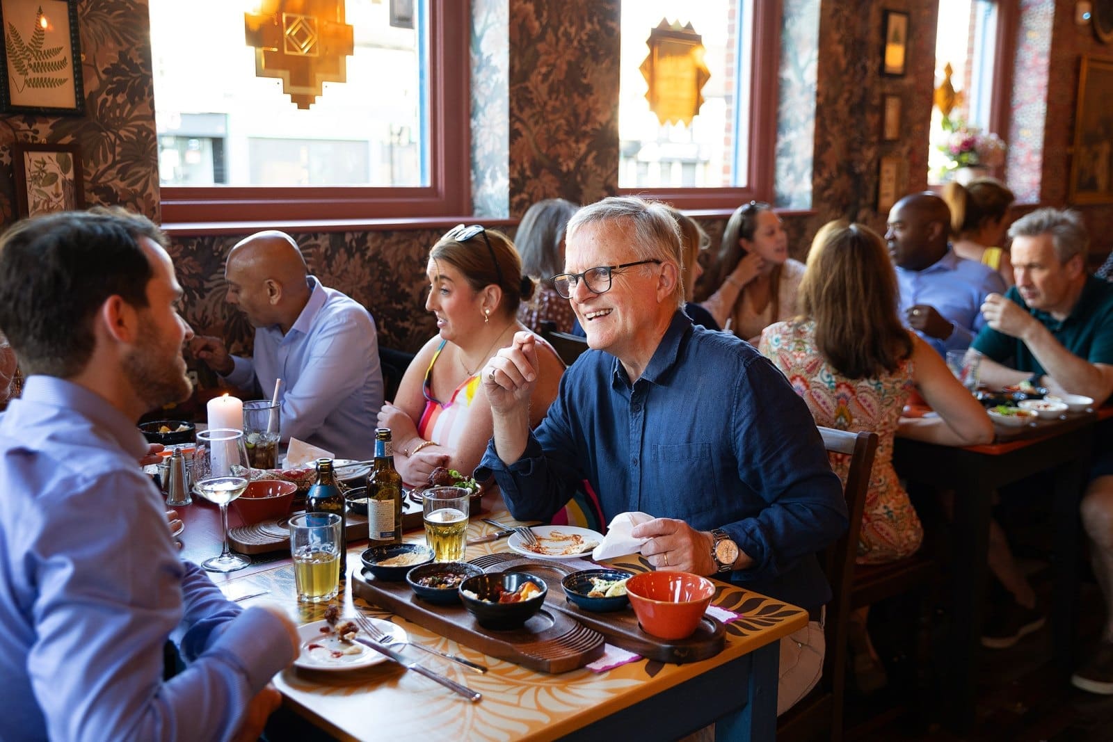 A group of people enjoy food and conversation at Carpino, a lively, warmly lit restaurant. Plates of food and drinks cover the tables, and everyone appears engaged and cheerful.