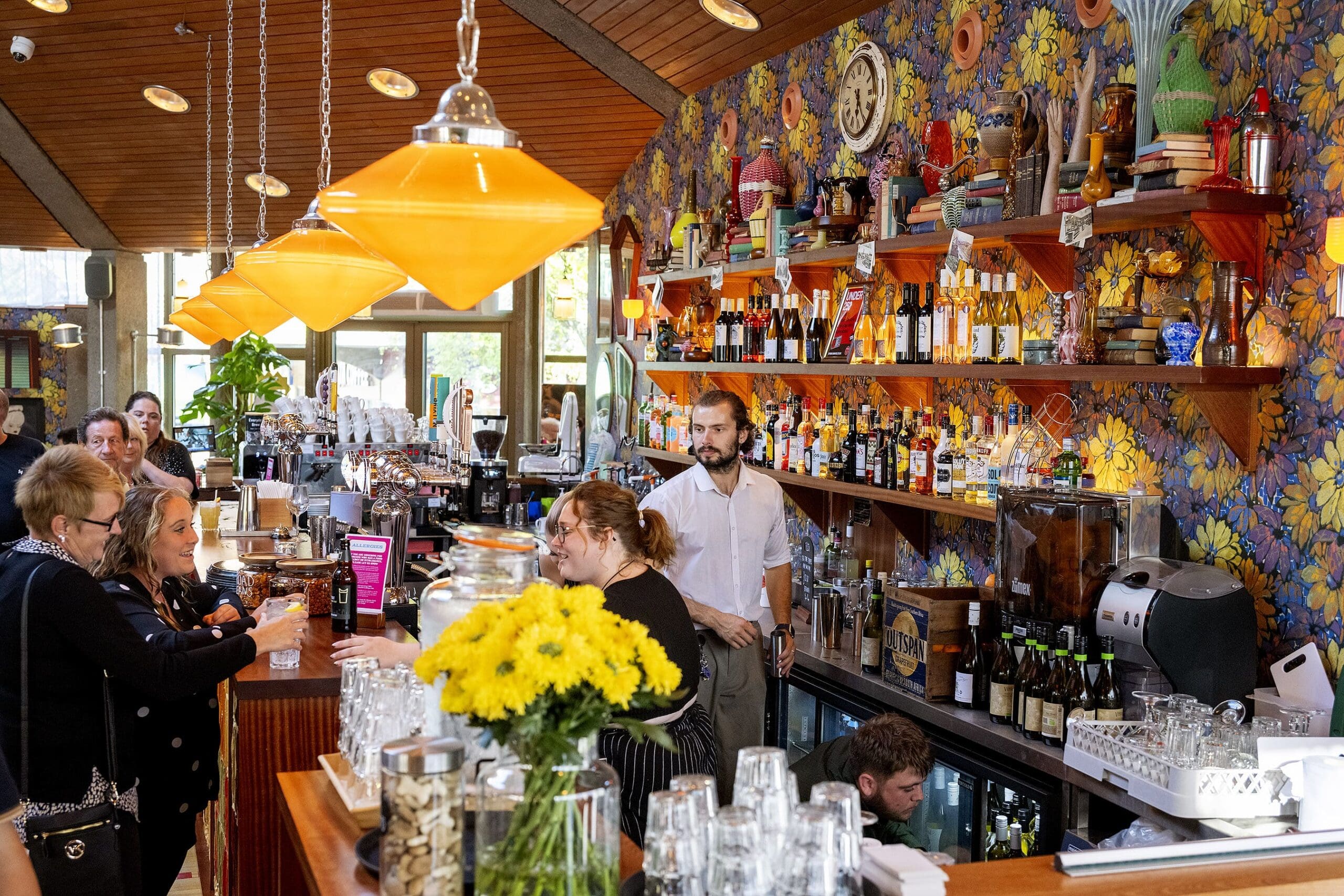 A lively café with colorful floral wallpaper, a busy bar with yellow hanging lights, people chatting and bartenders serving drinks, shelves filled with bottles, decor, and books like Livro, plus bright yellow flowers on the counter.