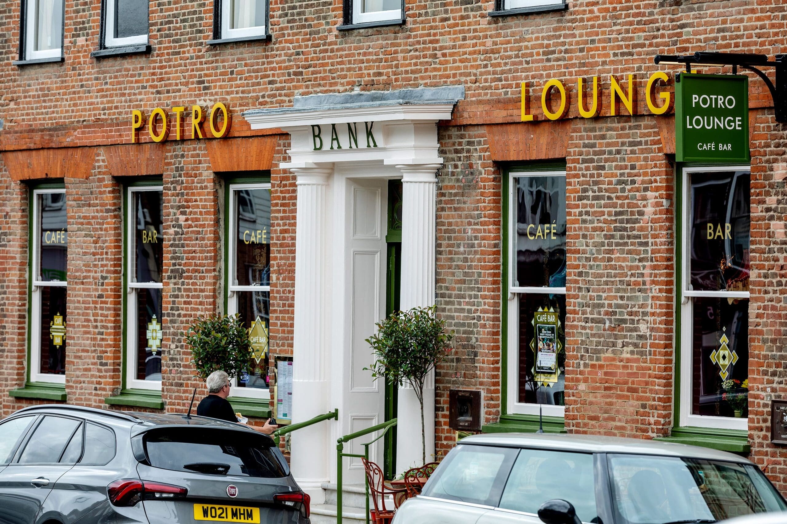 A brick building with “POTRO LOUNGE CAFÉ BAR” signs on the windows and “POTRO BANK” above the entrance stands out. Two small trees flank the door. Cars are parked in front, and a person waits by the sidewalk at Potro.