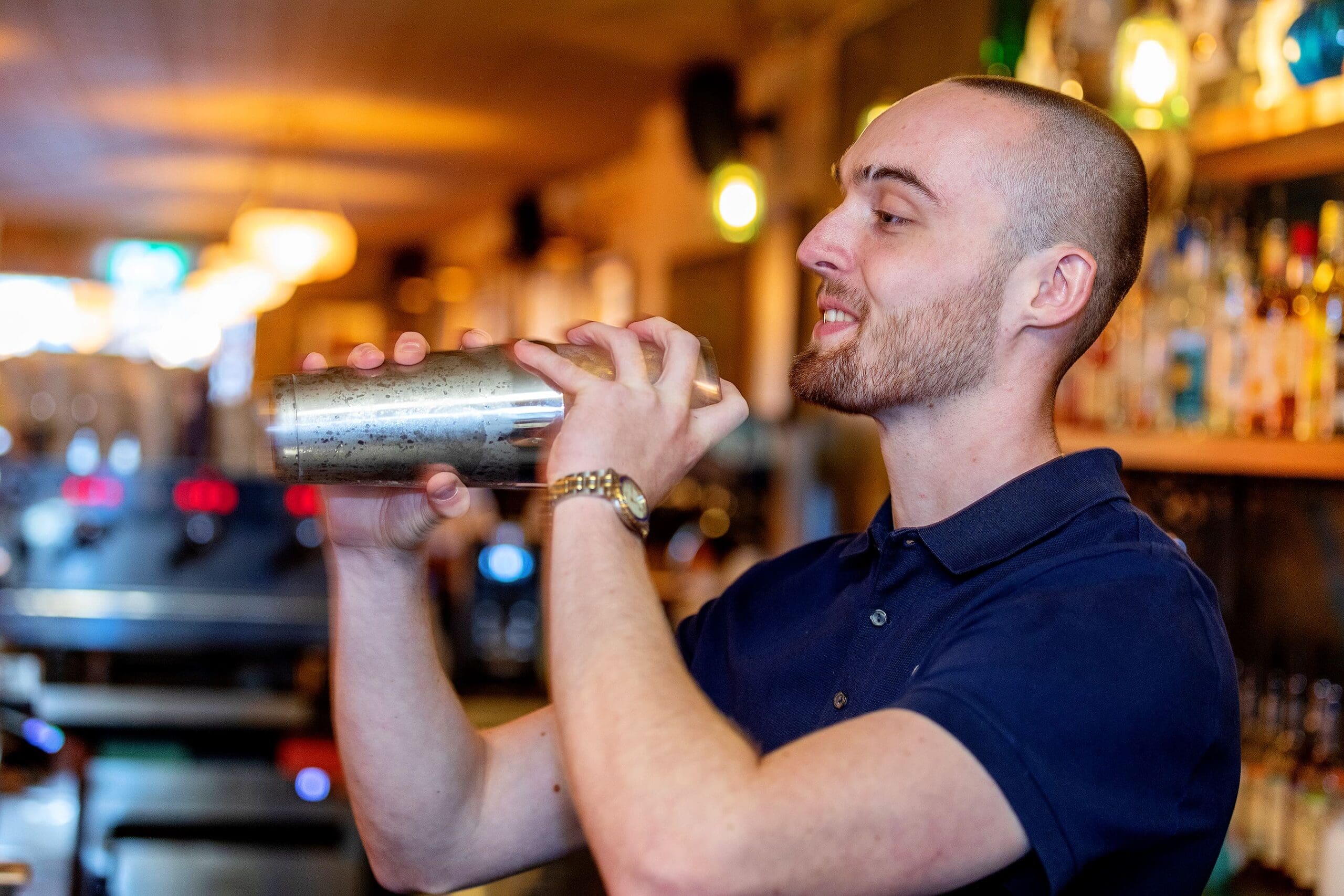 A smiling bartender in a navy blue shirt shakes a potro cocktail shaker behind a bar with shelves of liquor bottles in the background.