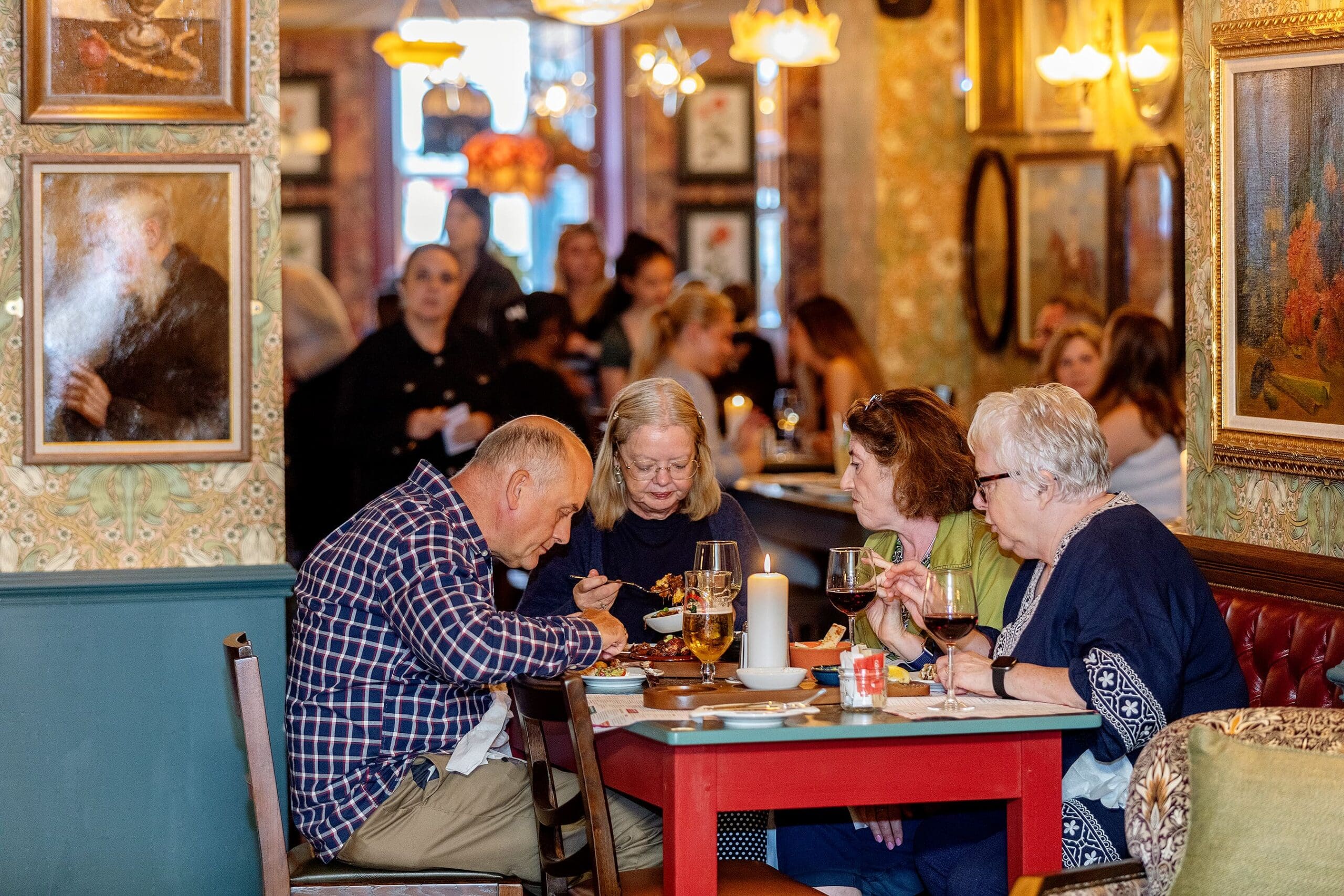 Four older adults sit at a restaurant table, eating, drinking wine, and sharing laughs over a potro dish, while other diners and staff move through the lively, warmly lit background filled with framed art on patterned walls.