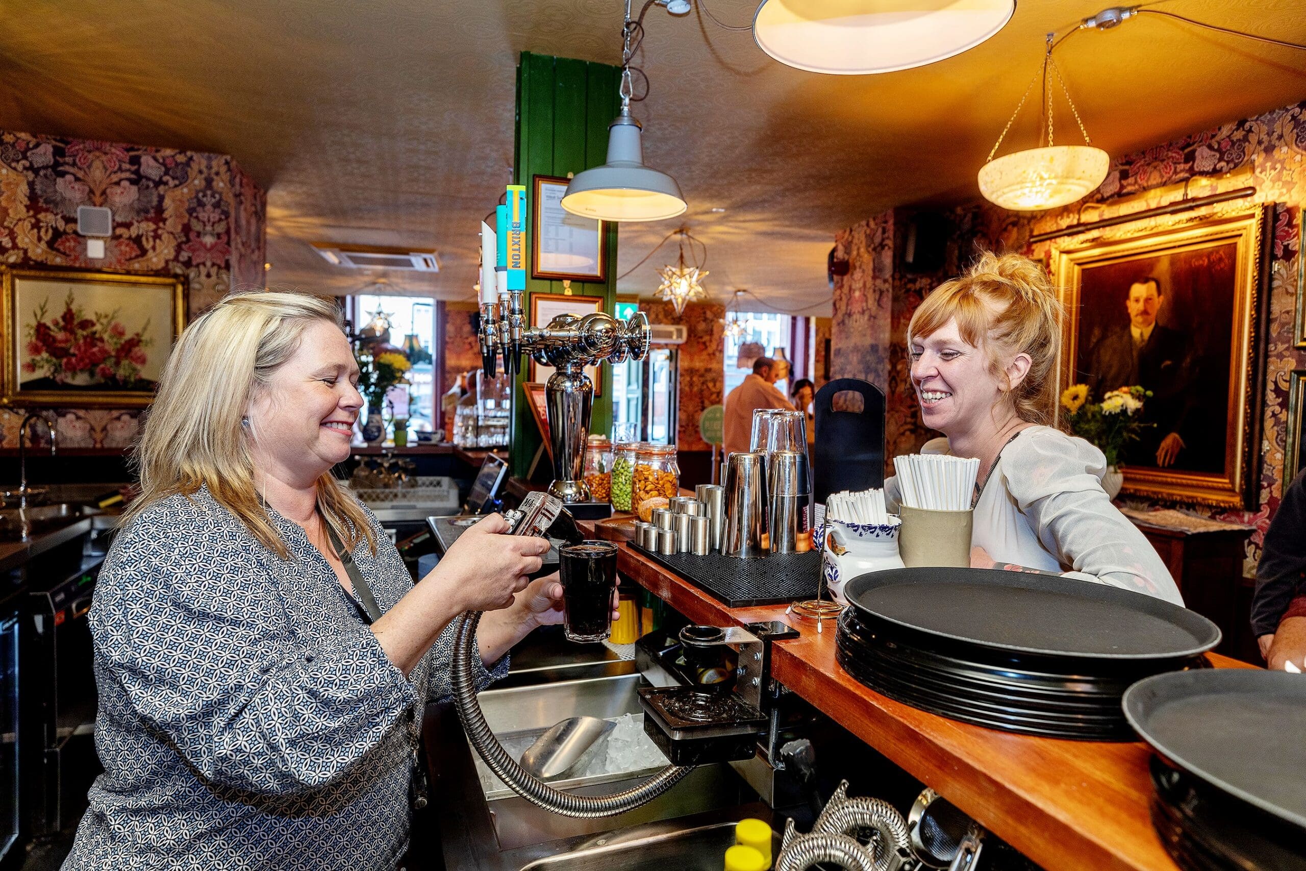 Two women smile at each other as one pours a drink from a potro tap behind the bar, surrounded by bar tools and glasses in a warmly lit pub with ornate wallpaper and framed paintings.