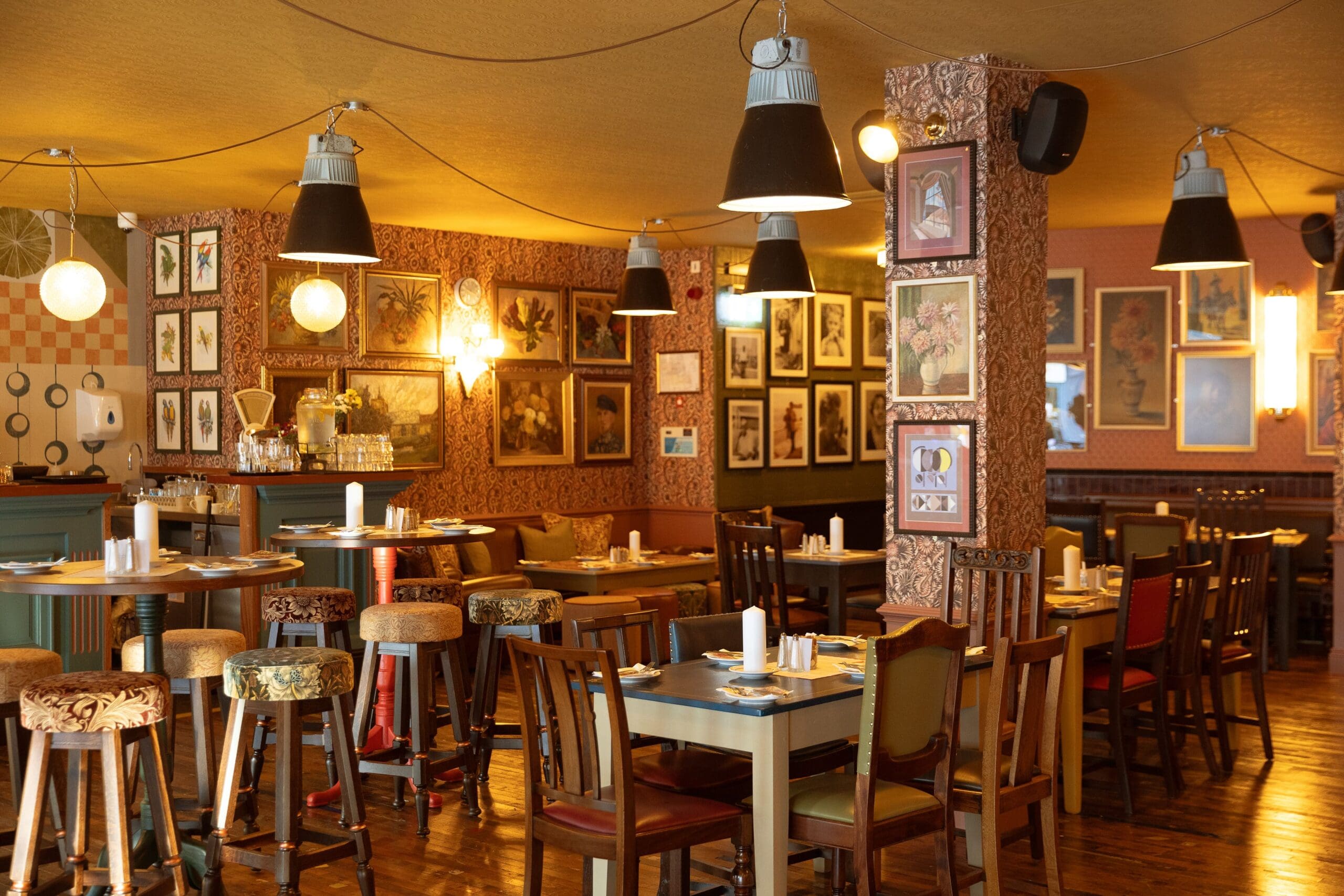A cozy restaurant interior features wooden tables and chairs, eclectic upholstered stools, warm lighting, and walls adorned with Maggio-inspired framed artworks and vintage decor. The tables are set for dining.