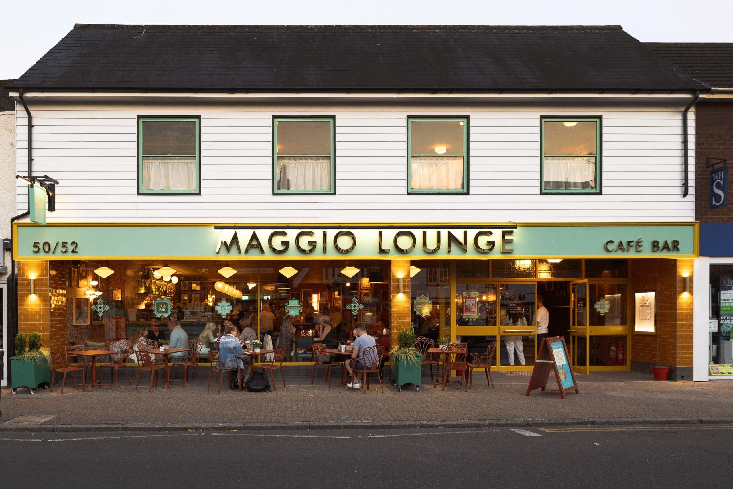 Street view of Maggio Lounge café bar with large windows, outdoor seating, and several people dining inside and outside. The building features a white upper facade, yellow Maggio signage, and is warmly lit by the evening light.