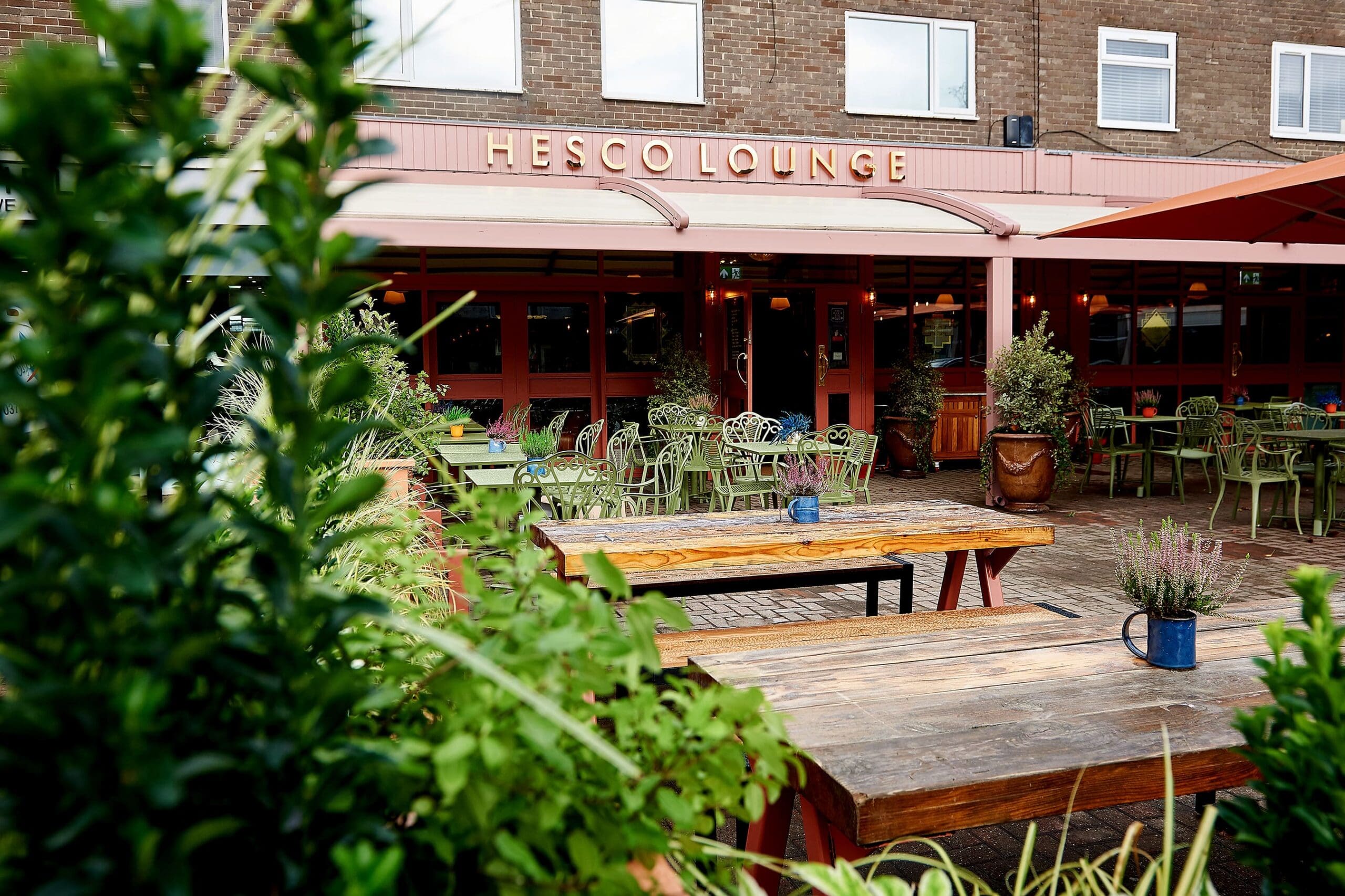 The outdoor seating area at Hesco Lounge features wooden tables, green chairs, and potted plants, set against the café's distinctive red and brown Hesco facade.