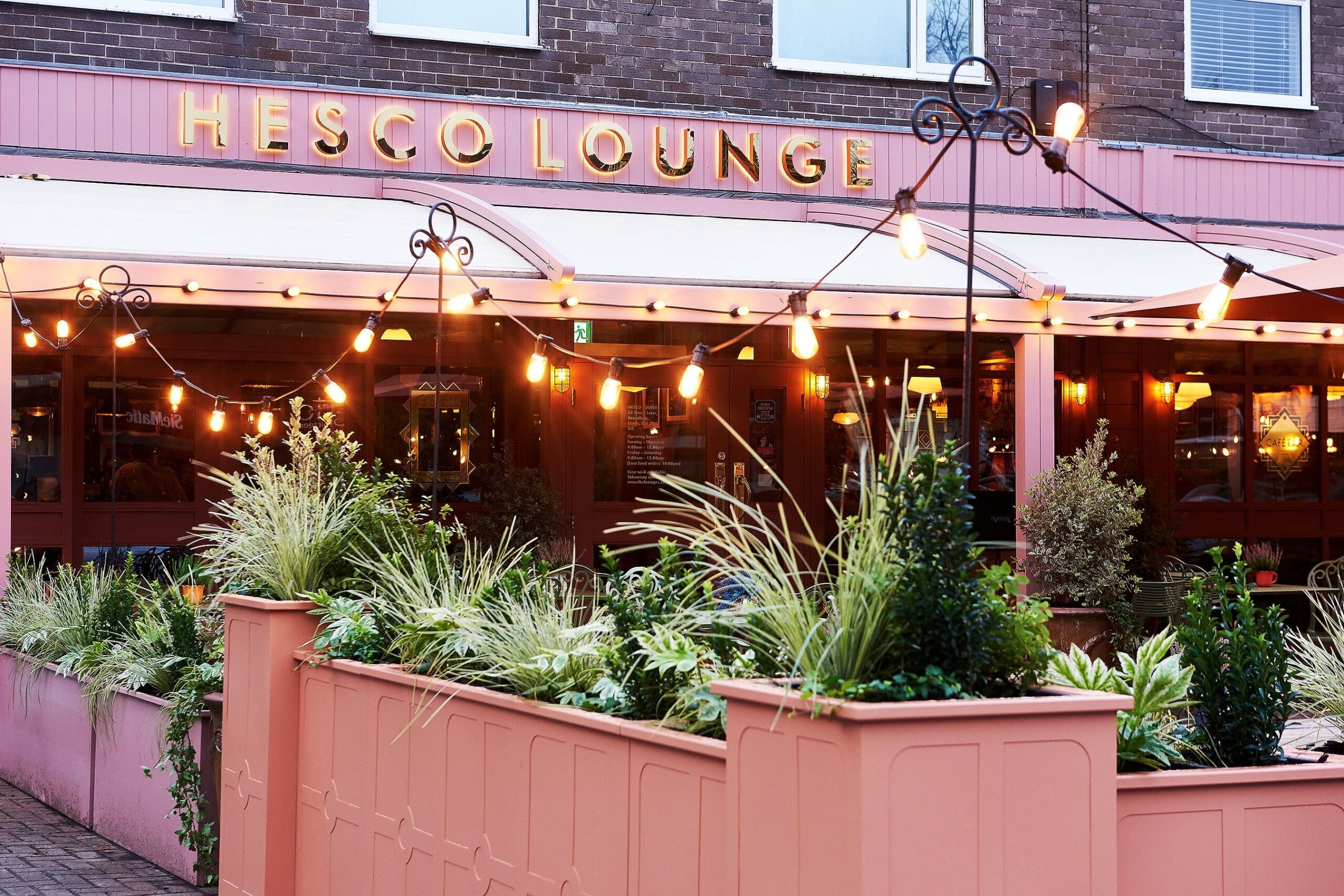 Outdoor view of Hesco Lounge café with string lights, large windows, and a pink patio filled with green plants in Hesco planter boxes. The atmosphere appears cozy and welcoming.