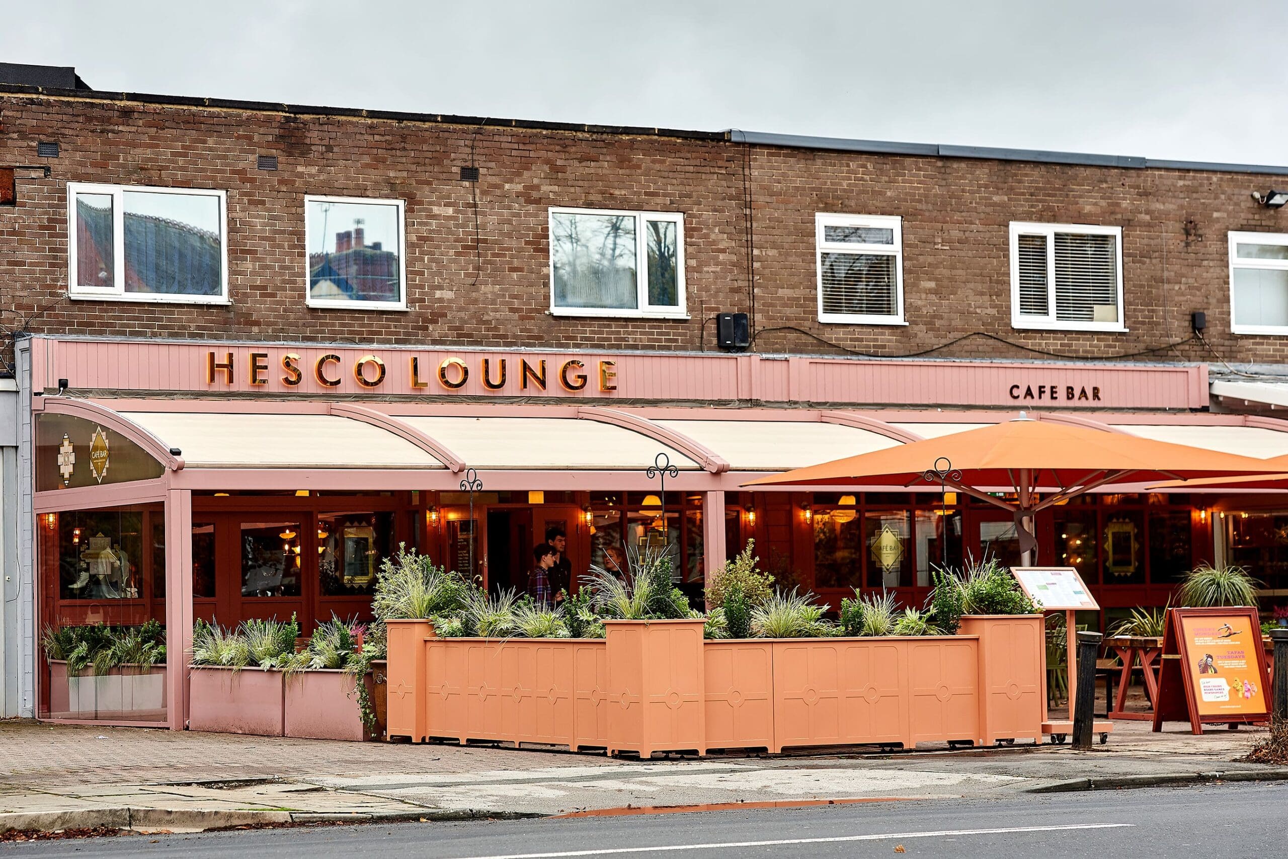 The exterior of Hesco Lounge, a vibrant café bar with a distinctive pink and orange facade, features large windows, outdoor seating under awnings and umbrellas, and potted plants lining the patio on a cloudy day.