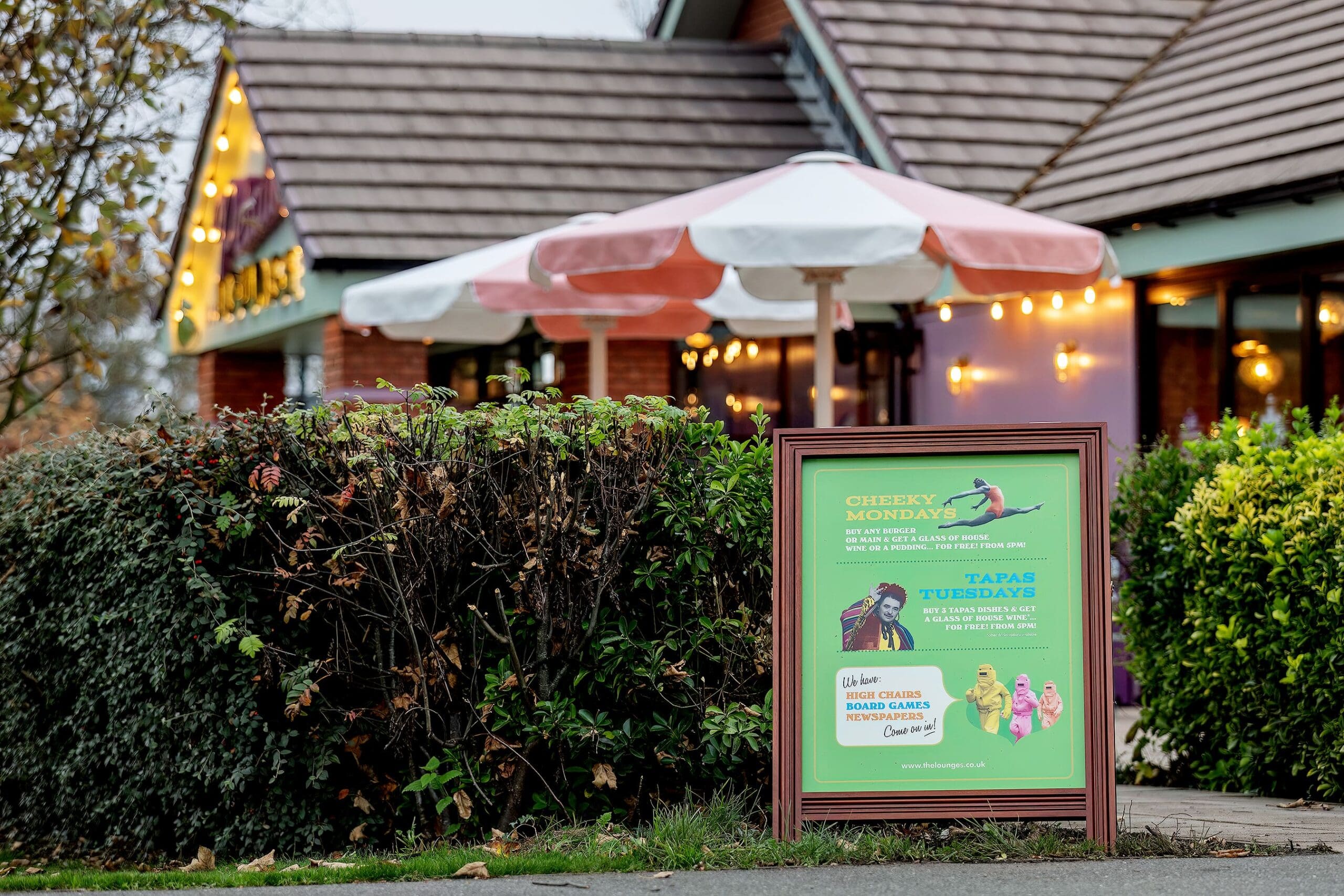 A restaurant with outdoor seating, petalo-pink and white umbrellas, and a large sign promoting Cheeky Mondays and Tapas Tuesdays offers next to green hedges. The atmosphere appears cozy and inviting.