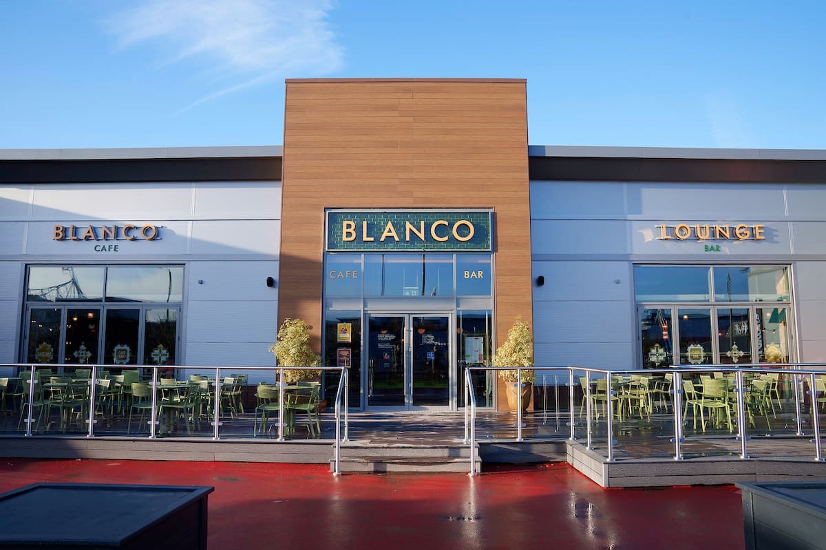 Exterior view of Blanco Café Bar & Lounge with large blanco signage, large windows, outdoor seating, modern wood and white facade, and clear blue sky above. Tables and chairs are arranged on the patio.