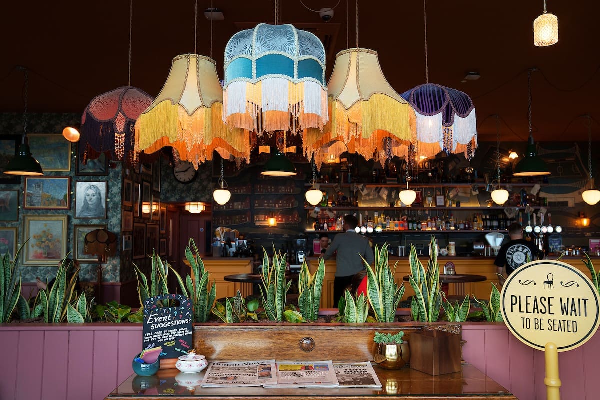 A cozy bar interior with vintage blanco lampshades hanging from the ceiling, potted plants on a wooden counter, framed art on the wall, and a “Please wait to be seated” sign in the foreground.