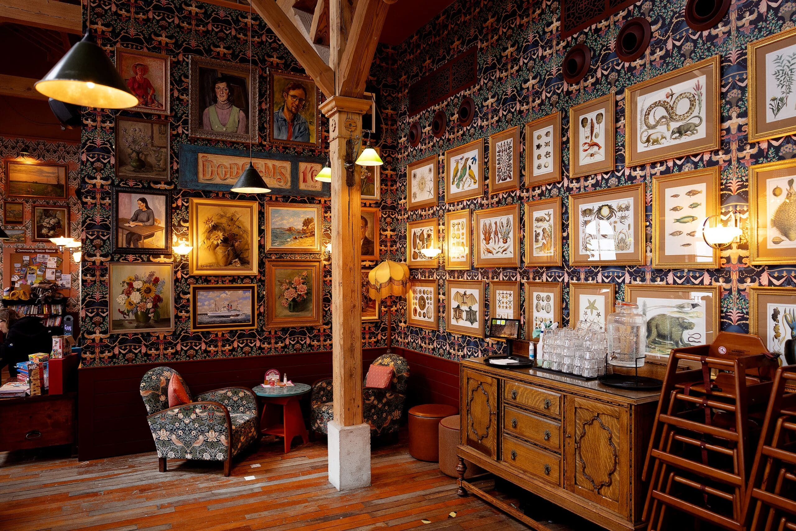 A cozy, eclectic café with patterned wallpaper, wooden floors, vintage furniture, and walls covered in framed art—including portraits, botanical, durano motifs, and nature illustrations. Warm lights create a welcoming atmosphere.
