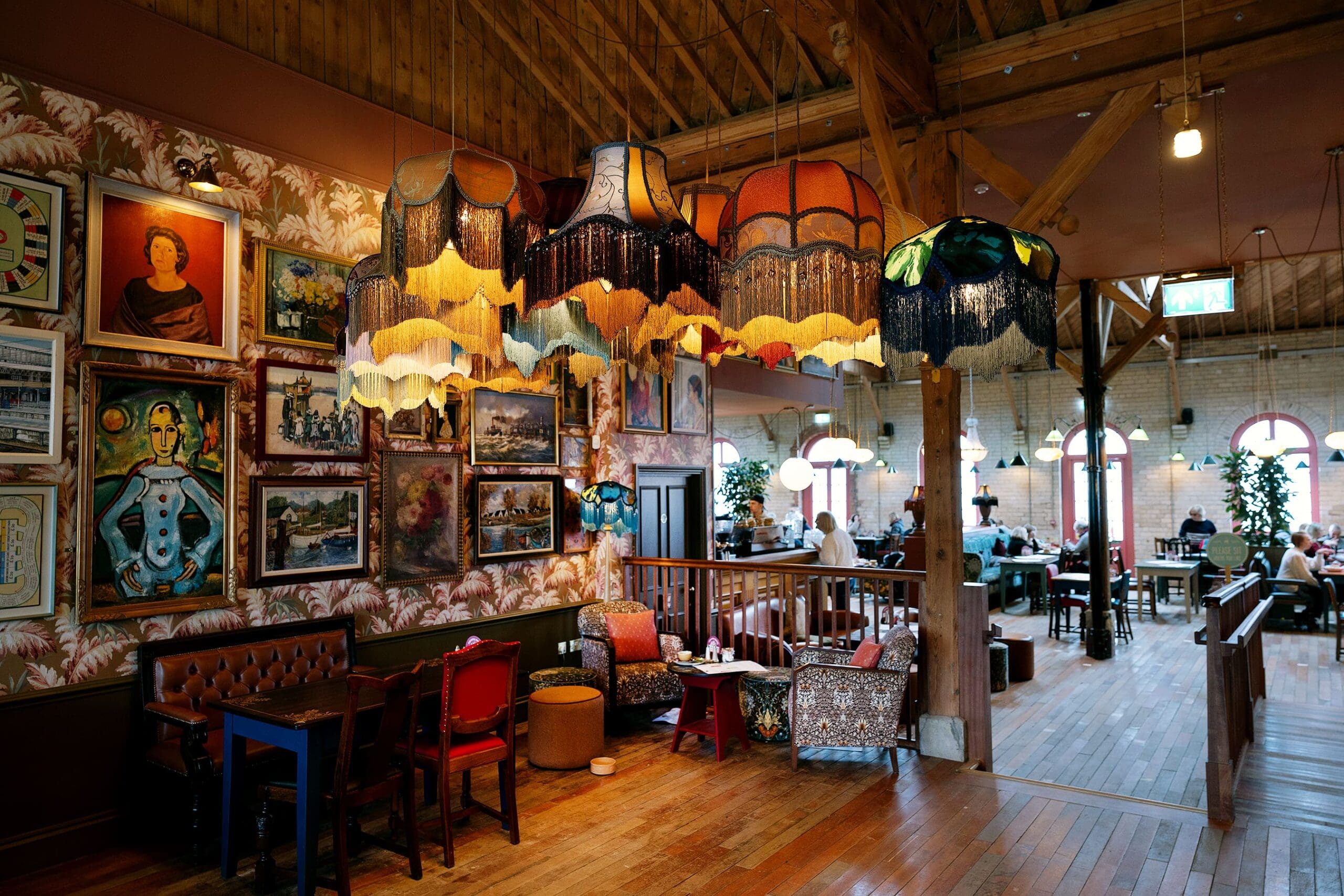 A cozy, eclectic cafe with vintage lampshades hanging from the ceiling, colorful paintings on patterned walls, wooden floors, mismatched furniture, and a touch of durano charm, with people seated comfortably in the background.