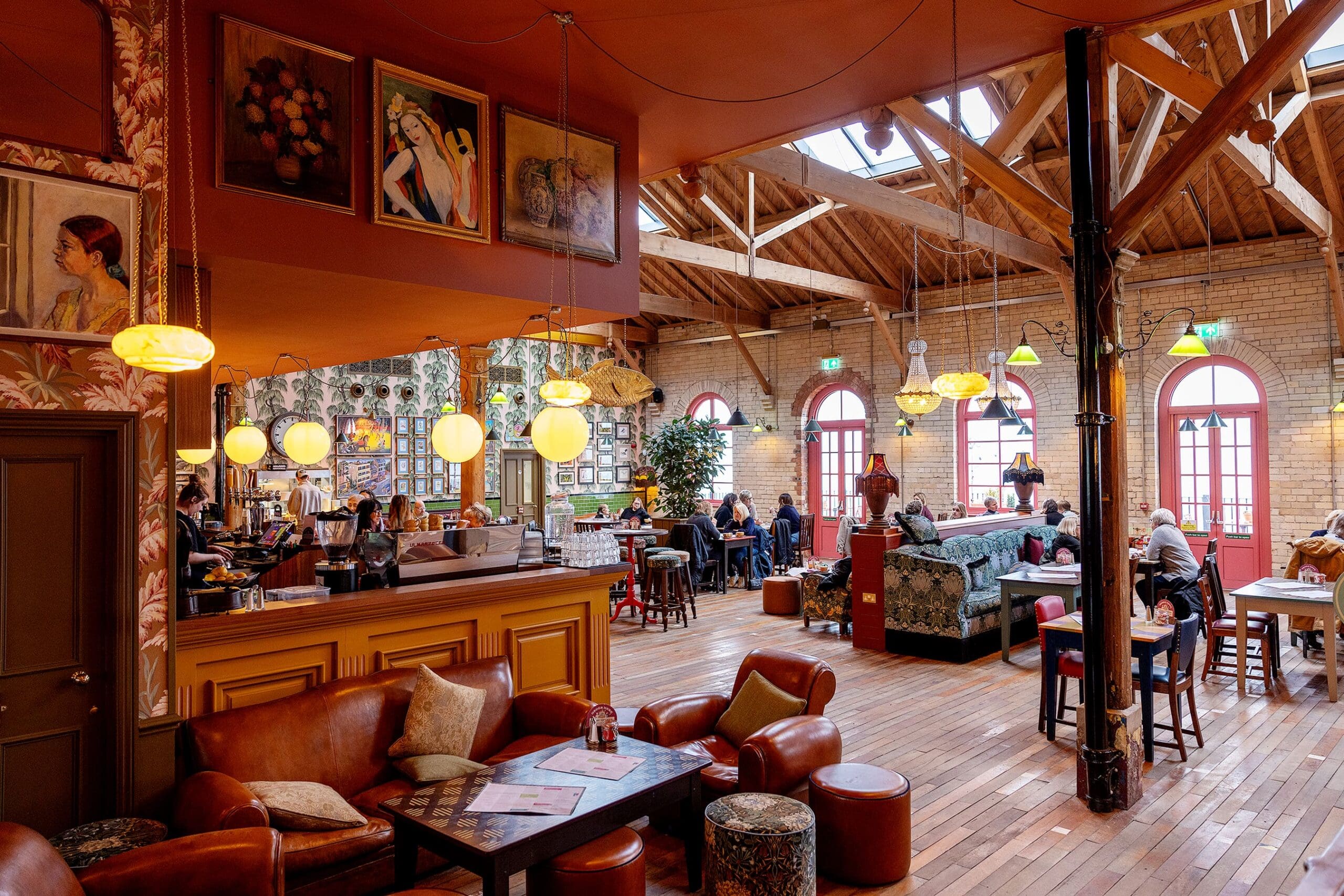 A spacious, warmly lit café with high wooden ceilings, vintage decor, hanging globe lights, durano leather seating, and people sitting at tables and booths enjoying drinks and conversation.
